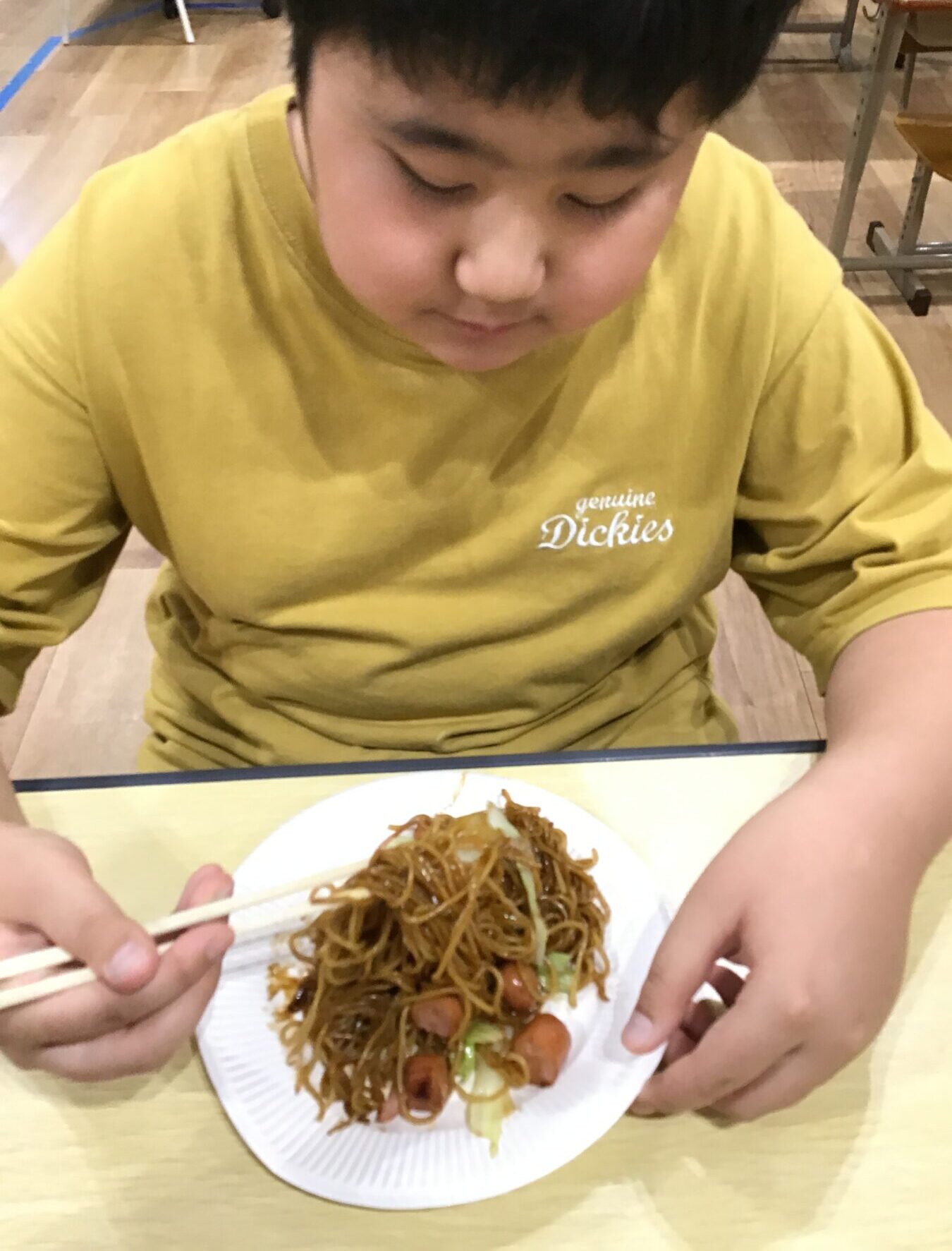 Person in a yellow shirt using chopsticks to eat stir-fried noodles from a white plate on a wooden table.