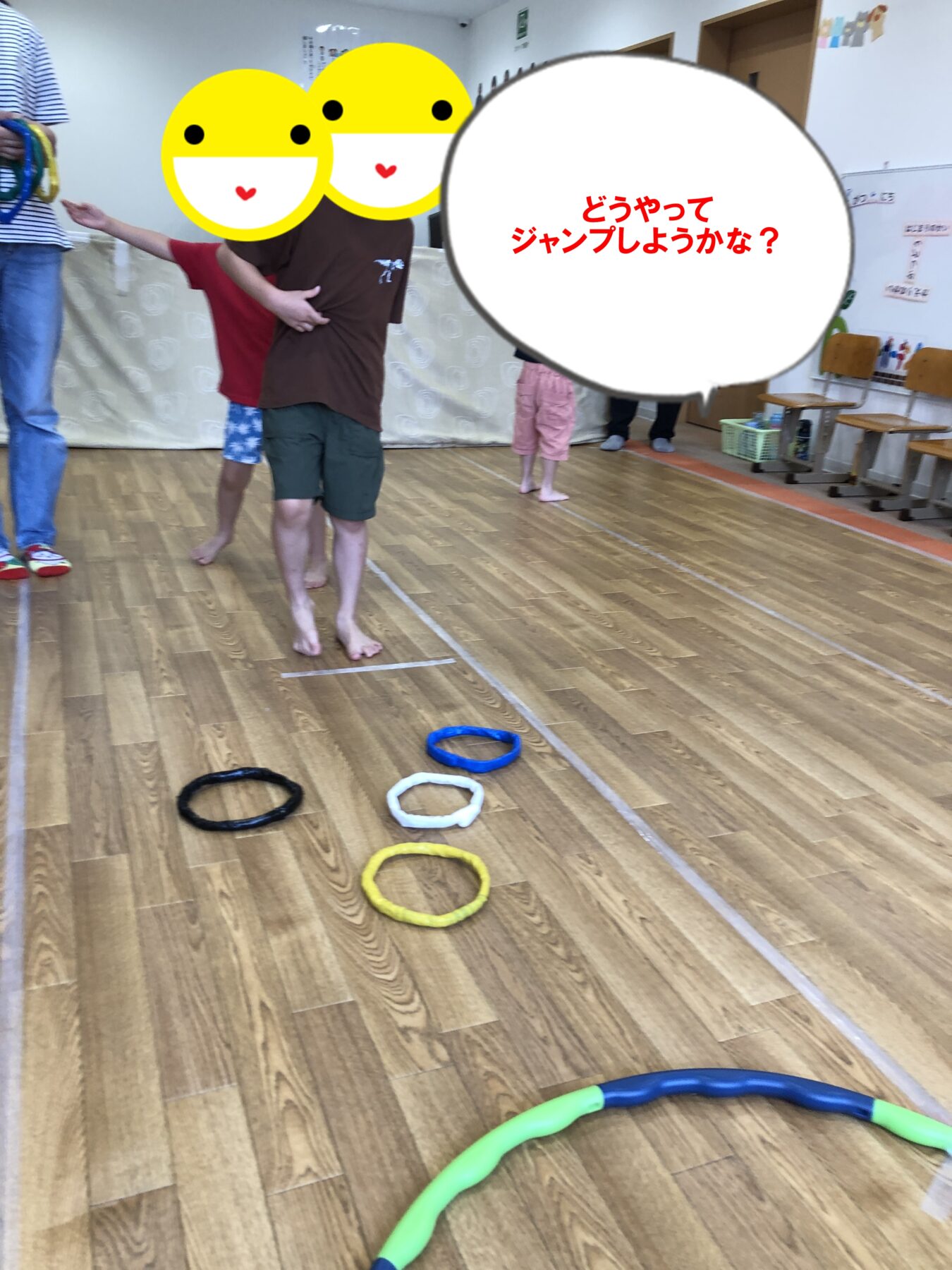 Children in a gym standing near hoop lines on the floor, preparing for a jump activity; speech bubble in Japanese asks, 'どうやってジャンプしようかな？'