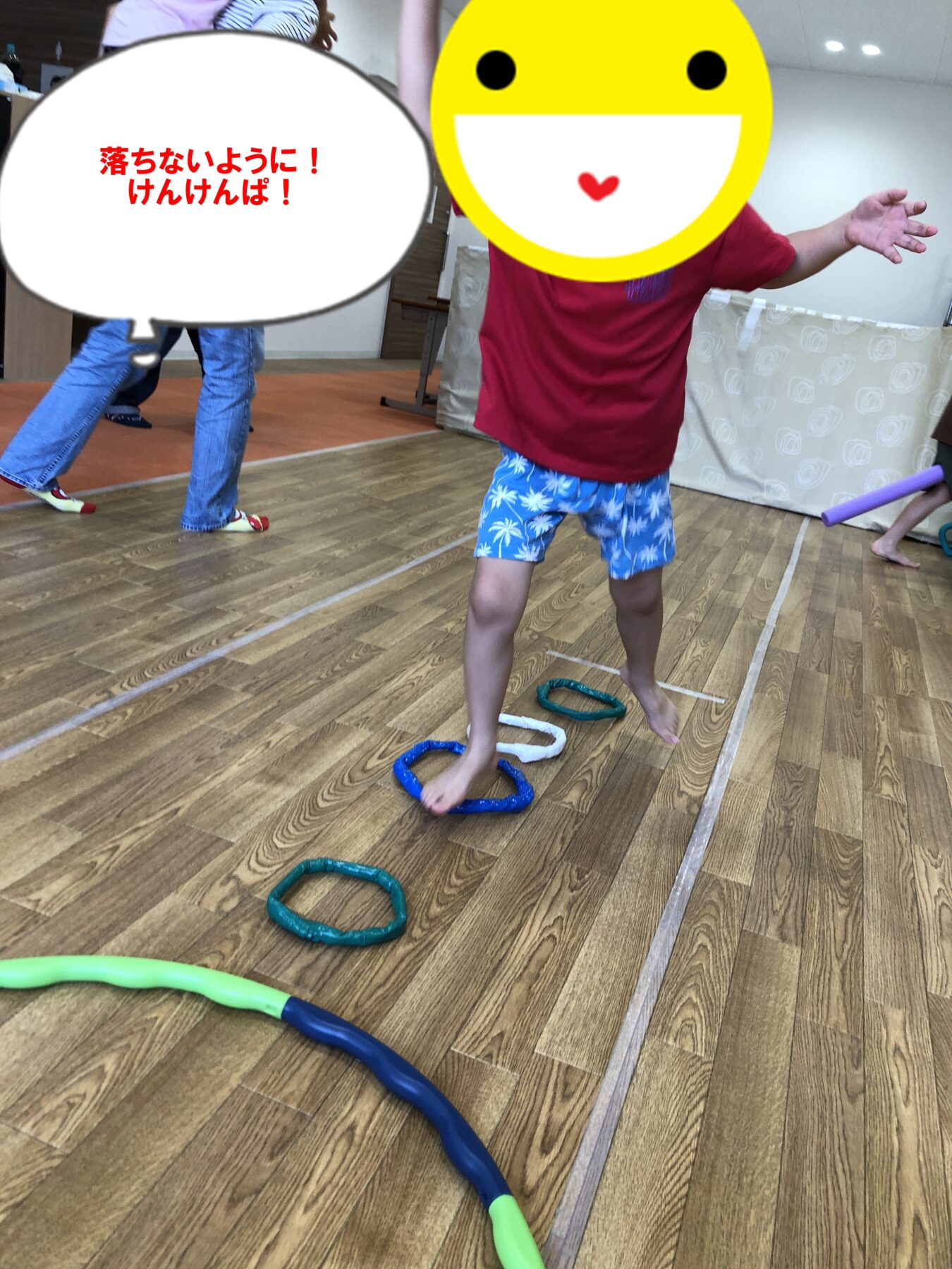 Child in a red shirt and blue shorts balances on circular hoops scattered on a wooden gym floor, with a large yellow smiley face overlaying the head.