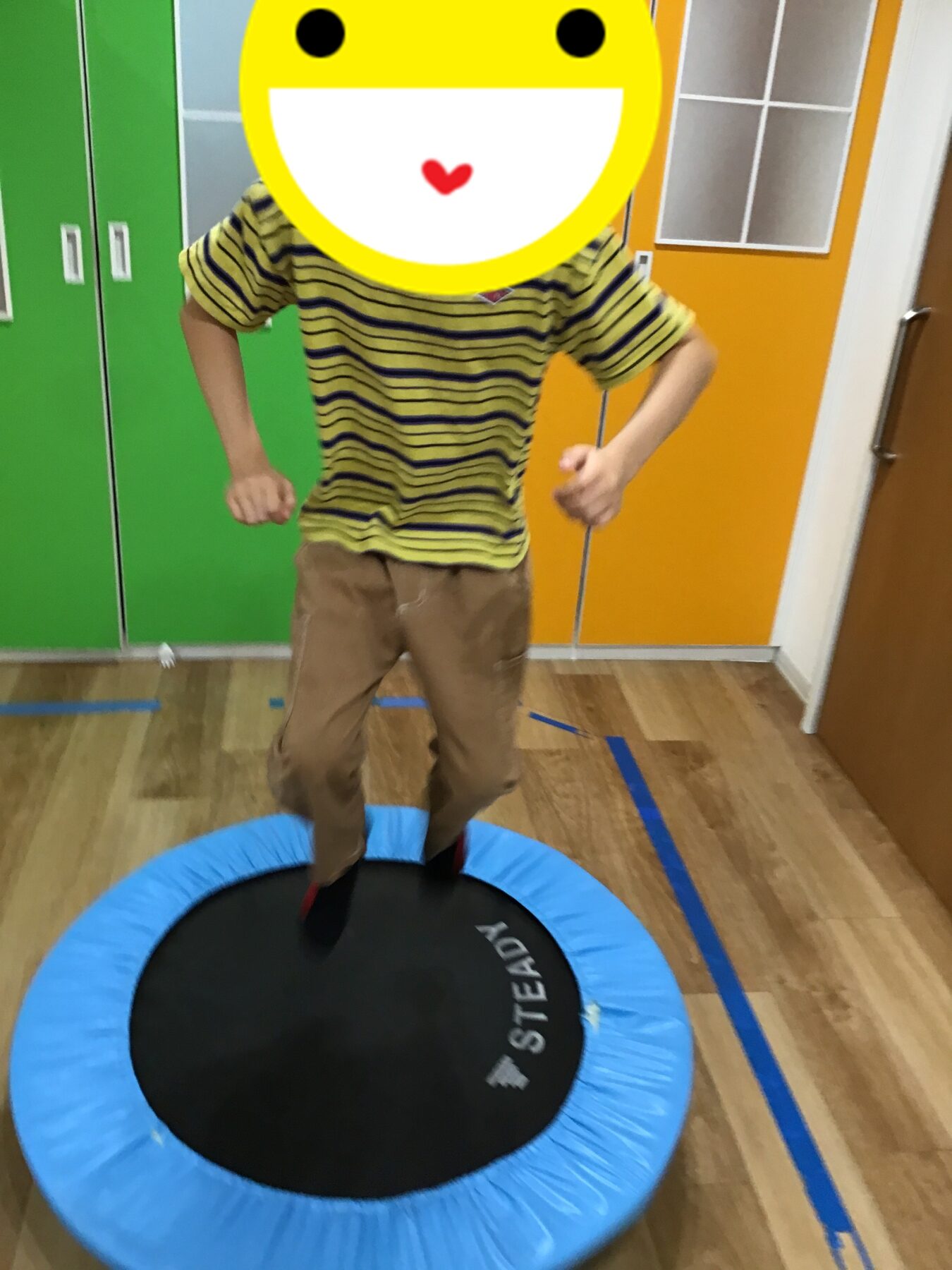 Child in a striped yellow shirt and brown pants jumping on a blue mini trampoline; face covered by a large yellow smiley emoji.