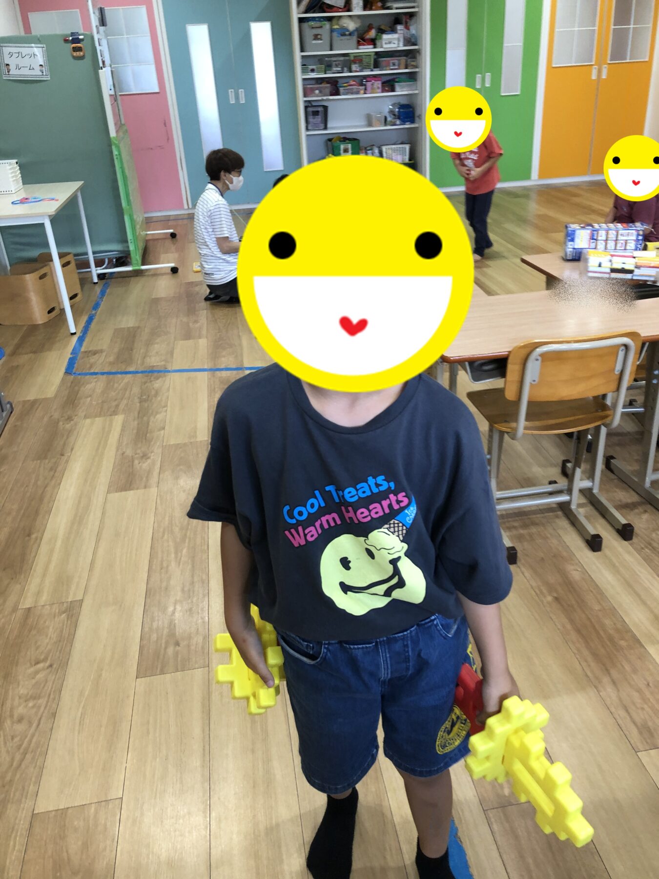 Child in a colorful classroom holding large yellow interlocking blocks, wearing a navy shirt reading 'Cool Treats, Warm Hearts'; shelves and bright doors in the background.