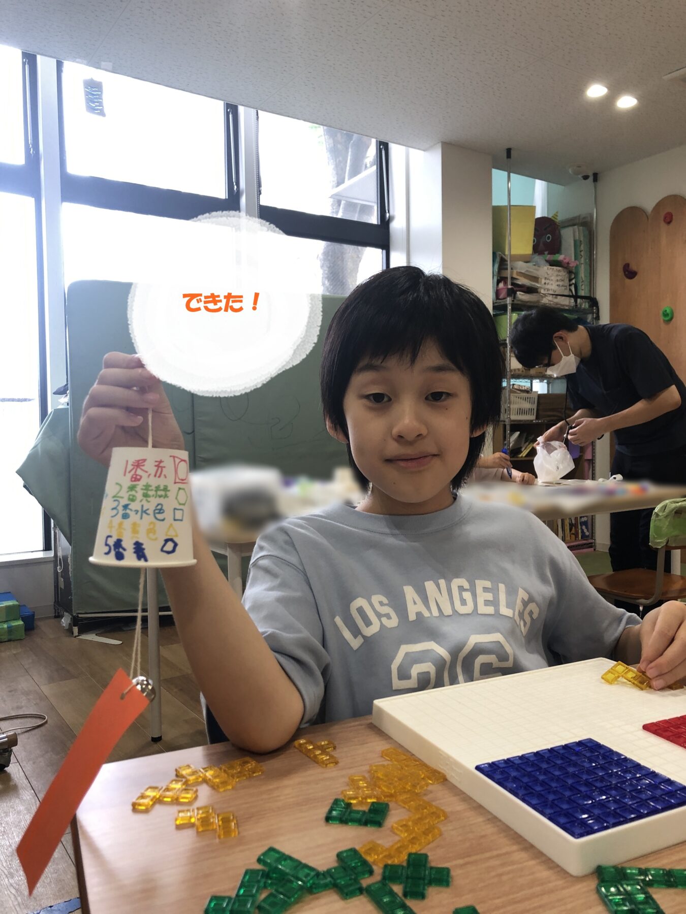 Young boy in a light blue shirt holds up a white circular hanging craft in a cluttered classroom. Building blocks in yellow and green sit on the table, with a teacher in the background.