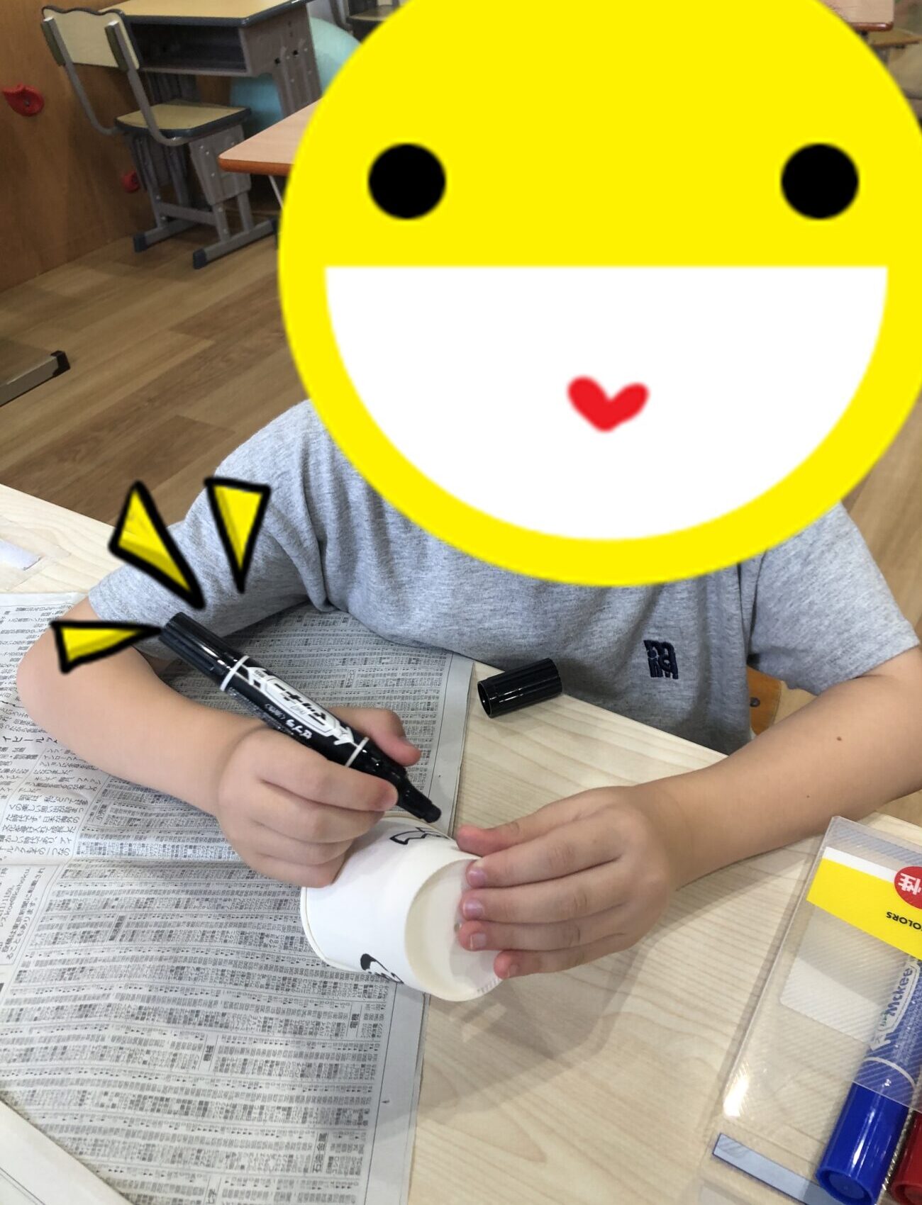 Child drawing on a paper cup with a black marker at a desk, classroom background, and a large yellow smiley-face graphic covering the child's face.