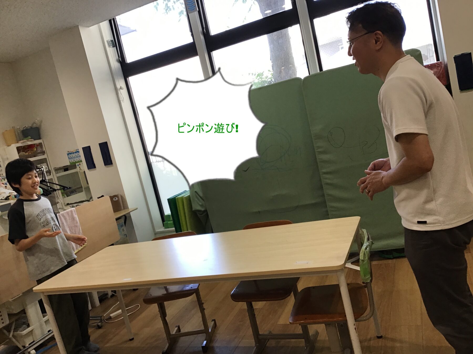 Young boy and an adult man stand in a classroom near a green partition, with a speech bubble reading 'ピンポン遊び!'.