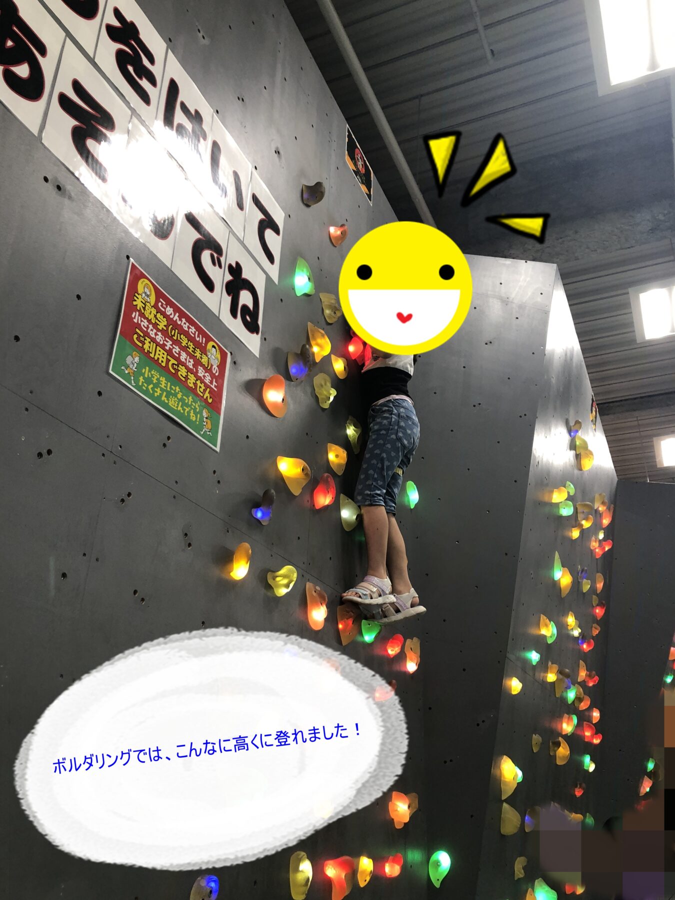 Child climbing an indoor gray climbing wall with colorful handholds and a wall sign nearby.