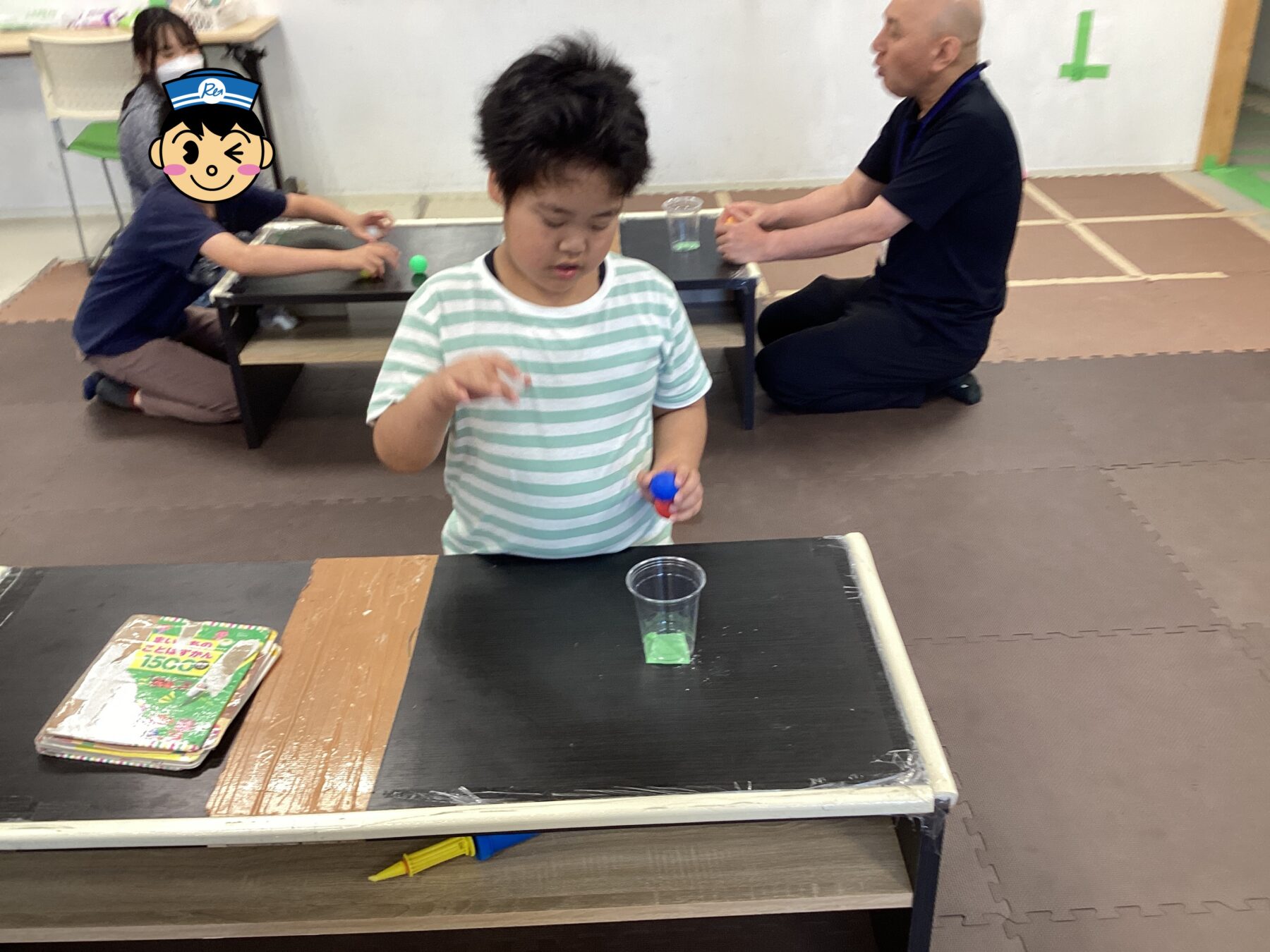 Child stands at a low table pouring green liquid into a plastic cup during a classroom activity, with another person seated at a separate table in the background.