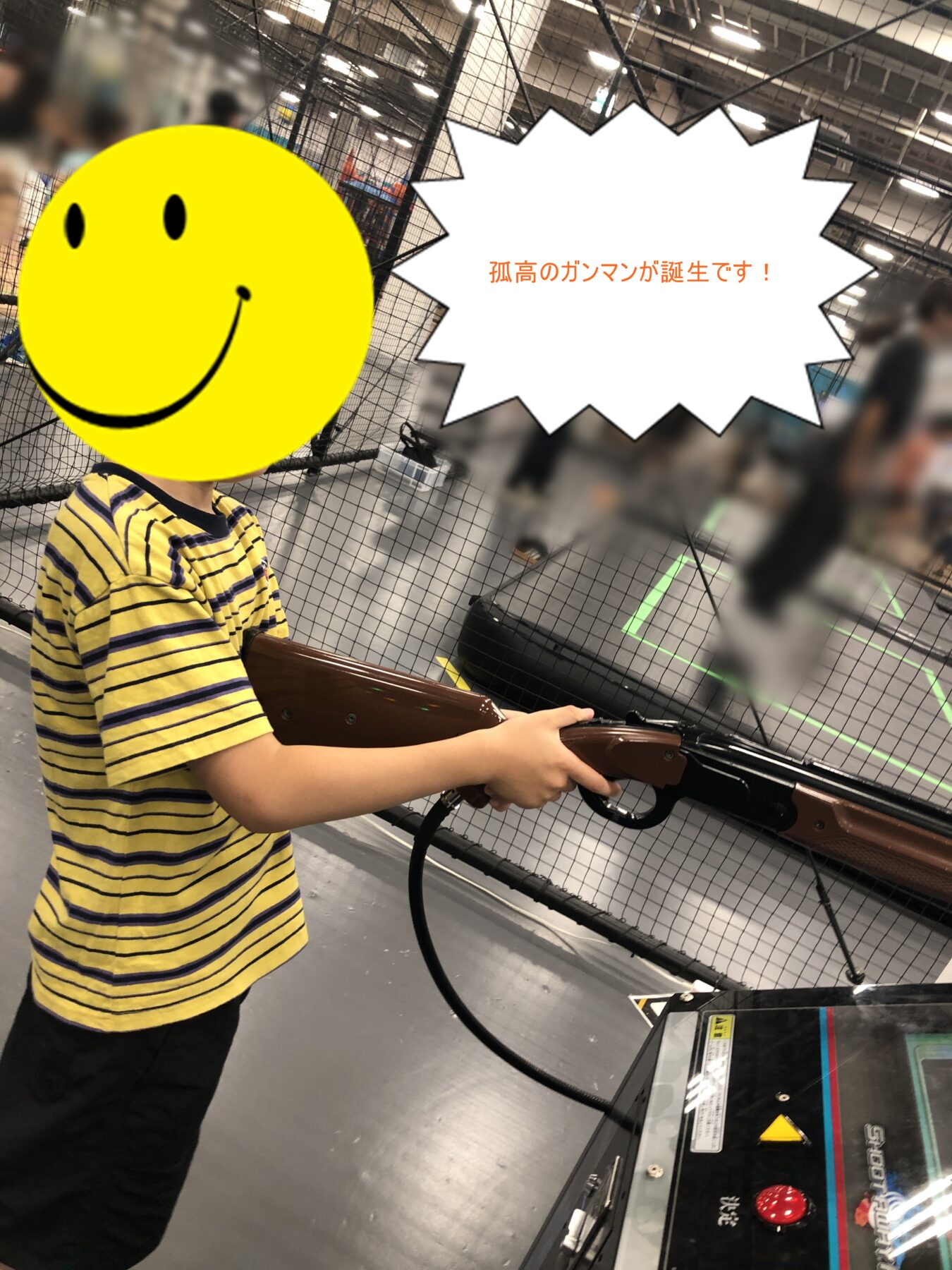 Child in a yellow striped shirt holds a brown arcade gun at a game venue; a large yellow smiley face sticker covers their face, with a comic speech bubble reading '孤高のガンマンが誕生です!'