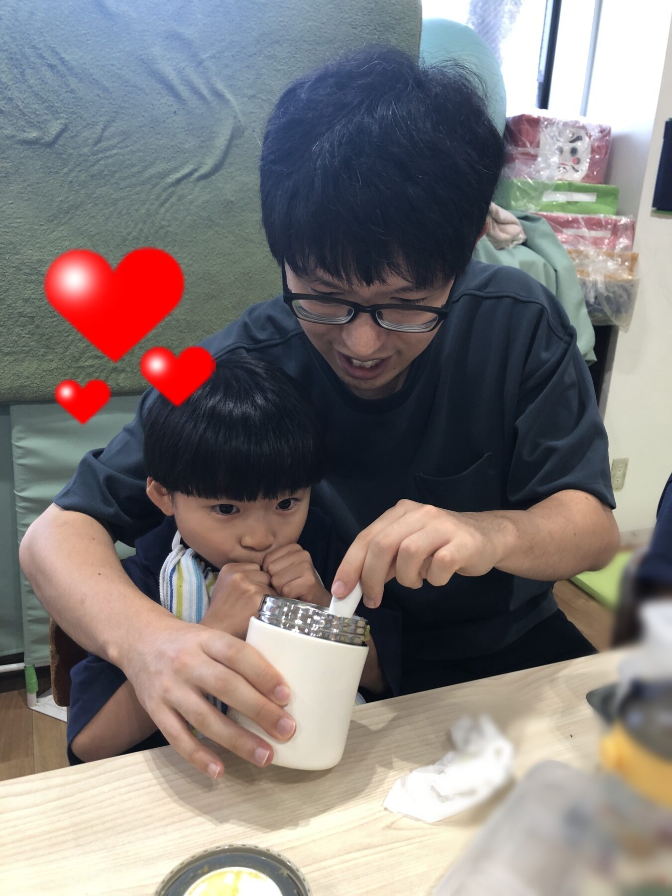 Man and child sitting together at a table, the man helping the child scoop a drink from a white tumbler; red hearts overlay convey affection.