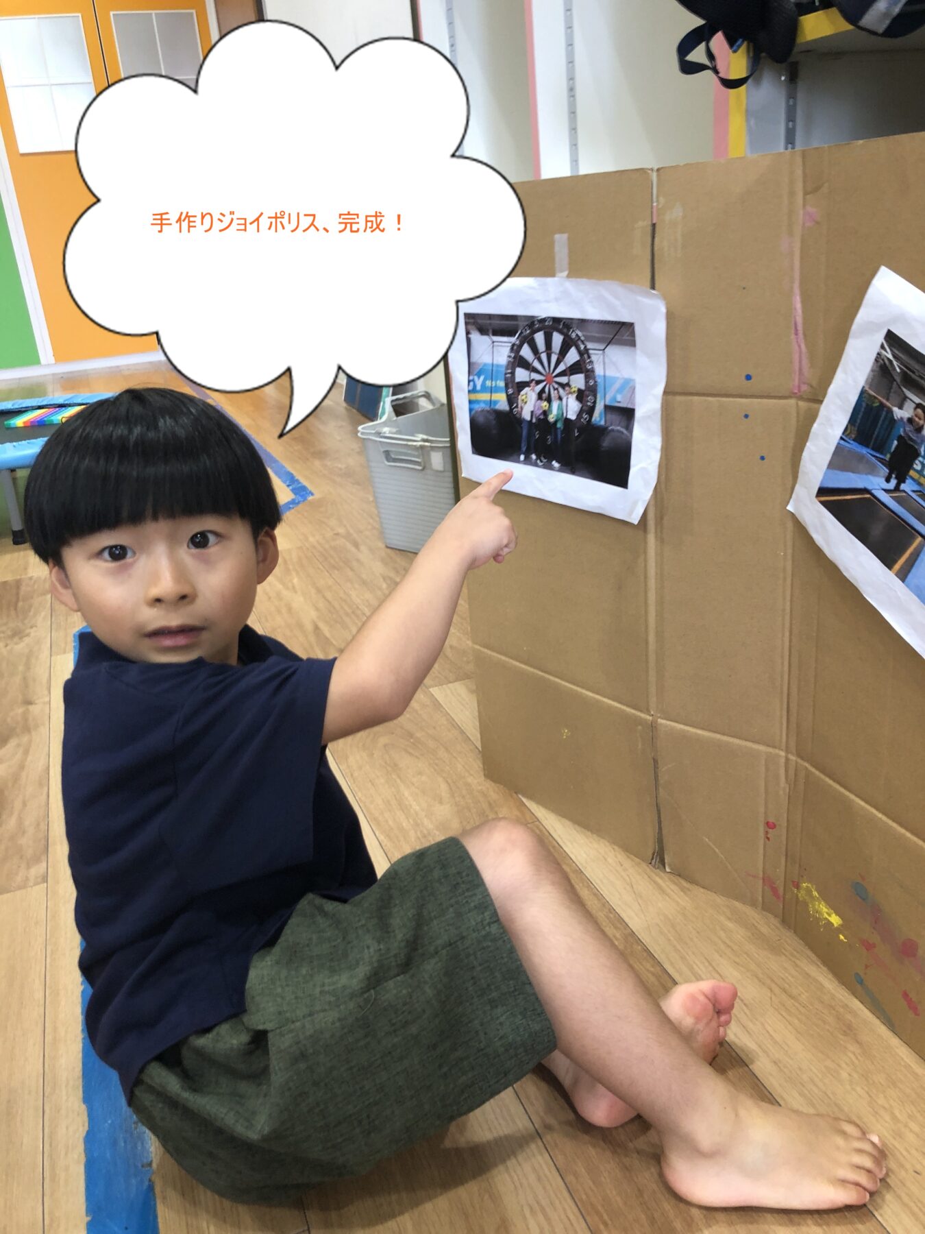 Boy sitting on a wooden floor points at a photo taped to a large cardboard display; speech bubble reads 手作りジョイポリス、完成! in Japanese.