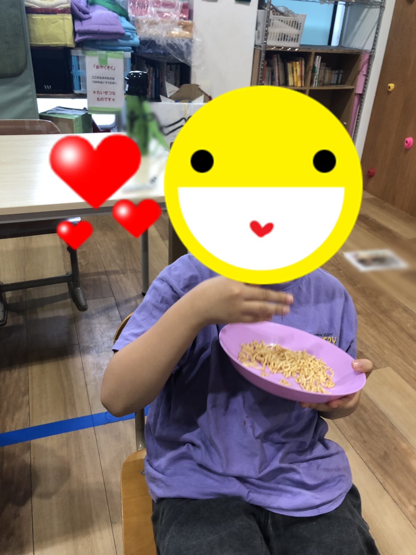 Child in a purple shirt sits at a table eating pasta from a pink bowl; a large yellow smiley emoji covers their face with heart graphics nearby.
