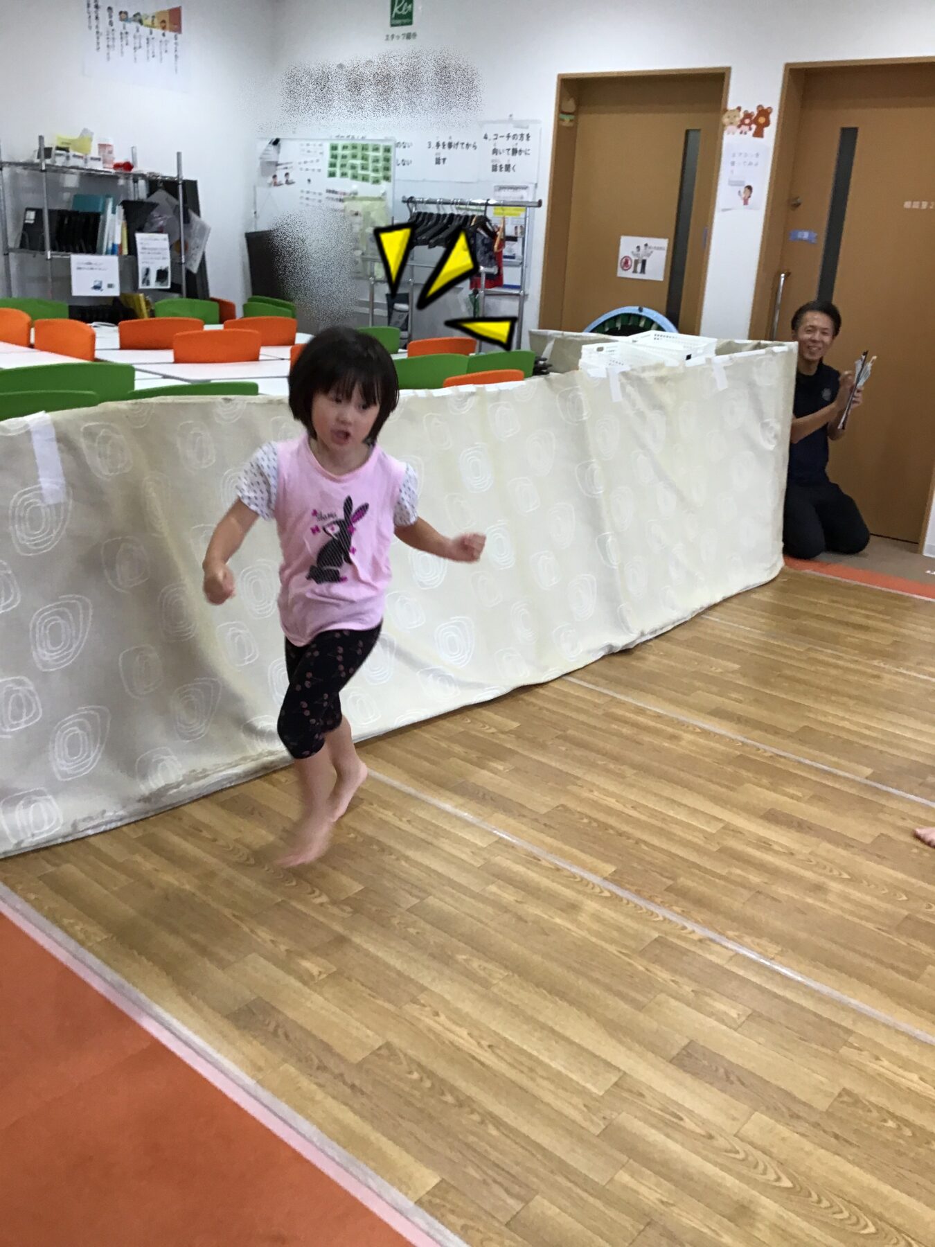 A young girl in a pink shirt runs barefoot across a gym-like room, with a colorful wall of chairs behind a white barrier.