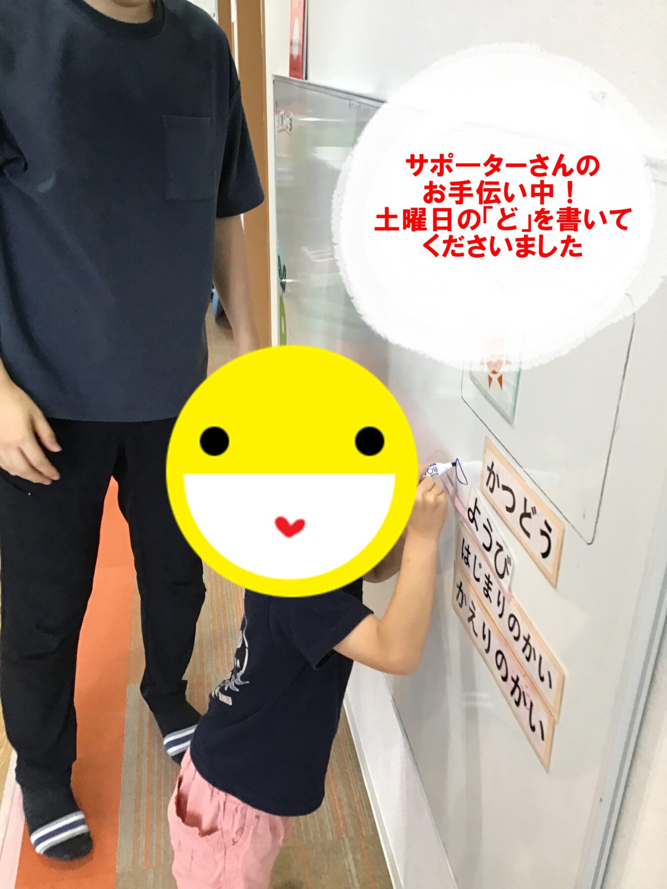 子どもが大人の見守る中、壁の掲示板に日本語の文字を書いている。