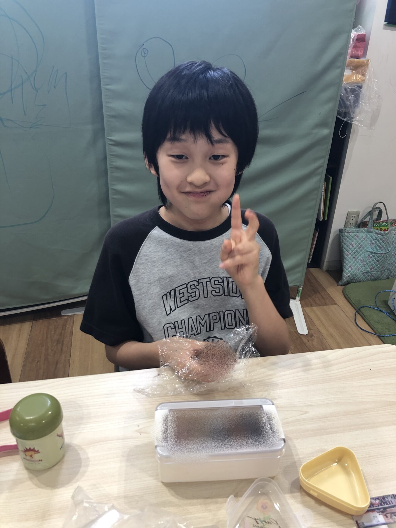 Smiling child with dark hair making a peace sign at a table with crafting supplies nearby in a classroom setting.