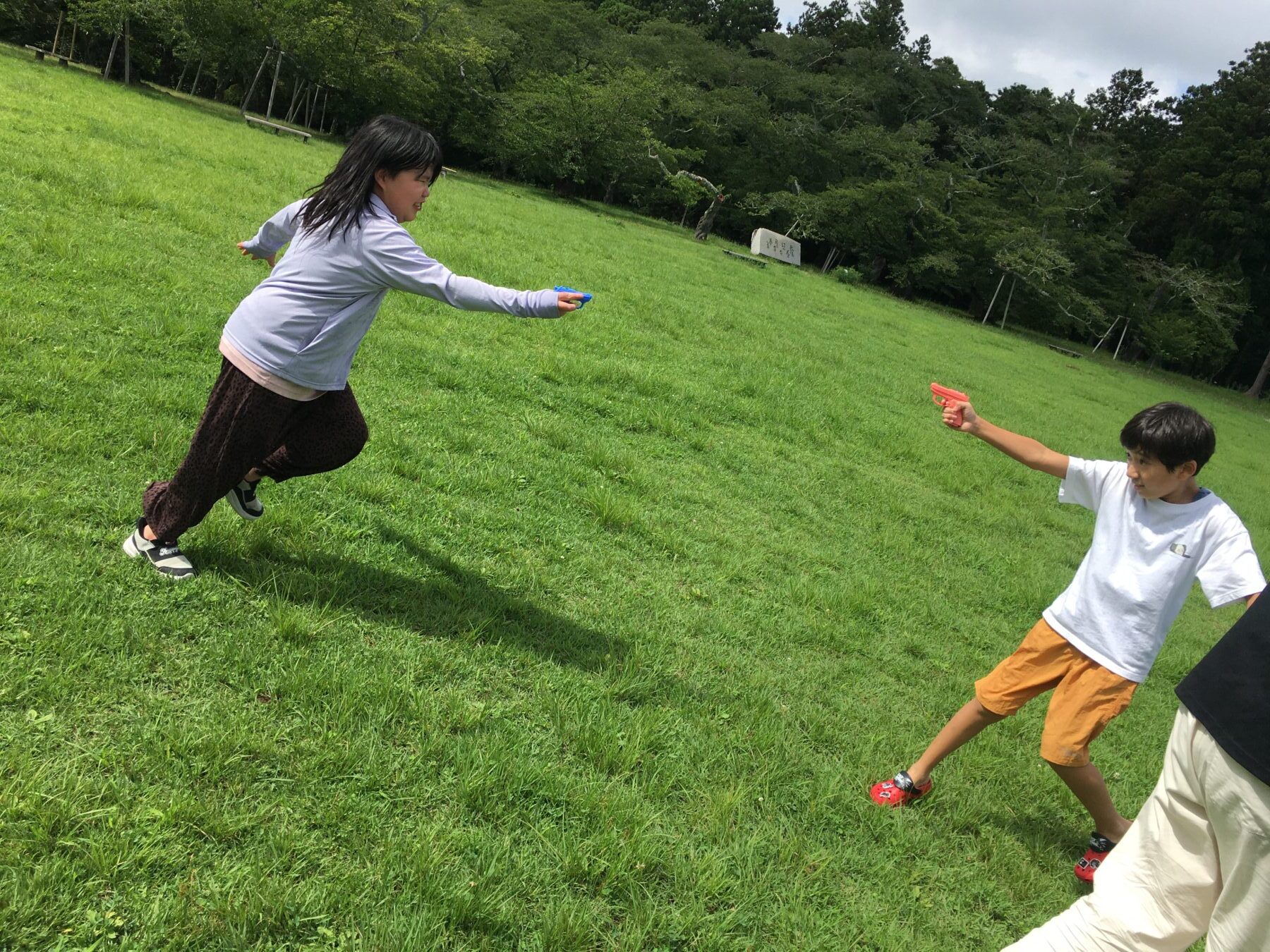 Two children playing catch on a grassy park lawn, tossing small red discs toward each other.