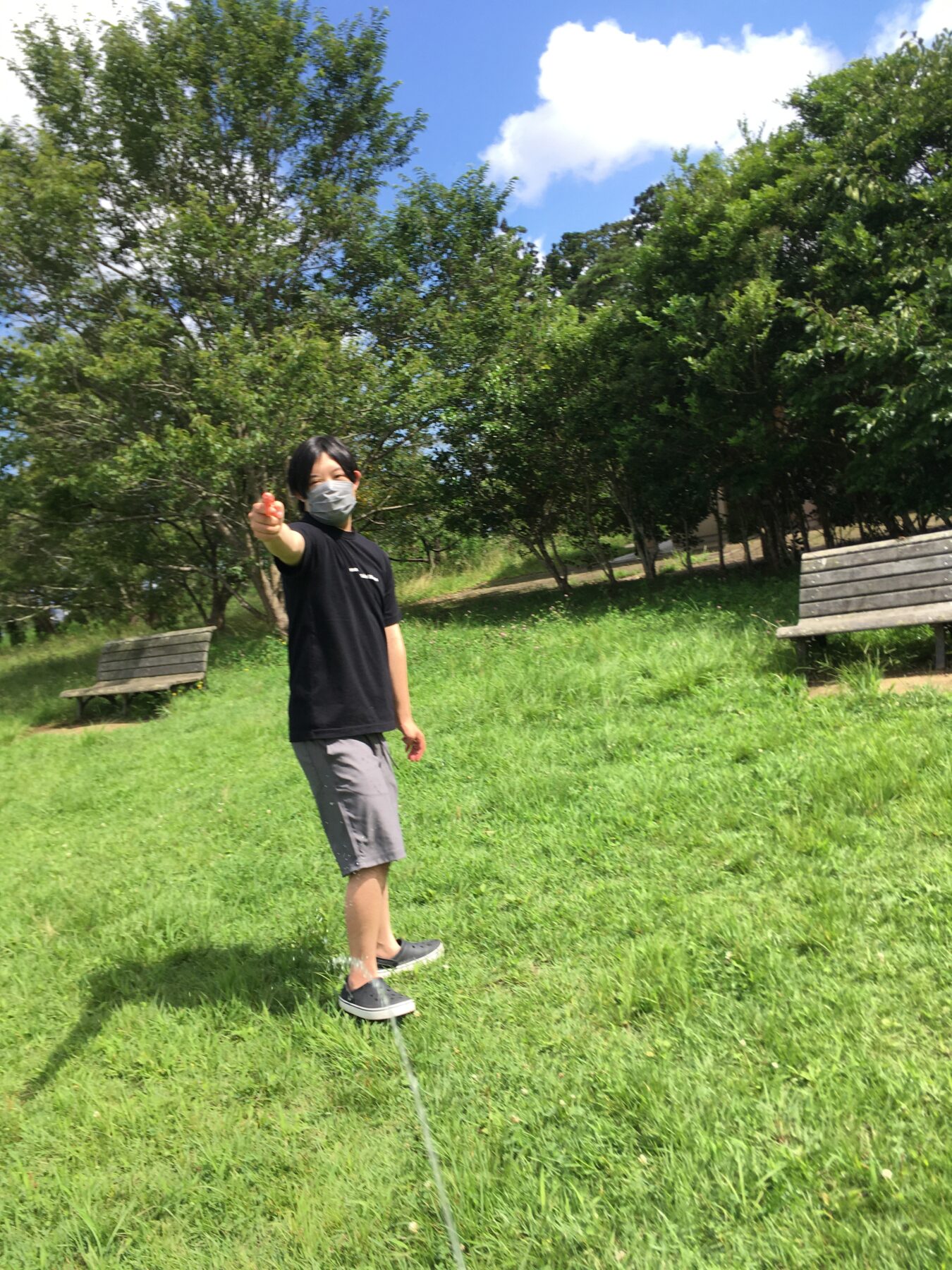 Person wearing a face mask points toward the camera in a sunny park with green grass, trees, and wooden benches in the background.