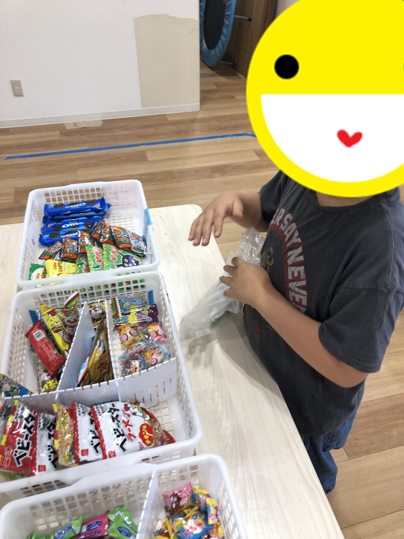 Child standing at a table sorting through baskets of assorted candy packets, some colorful wrappers visible in white baskets on a light wood table.