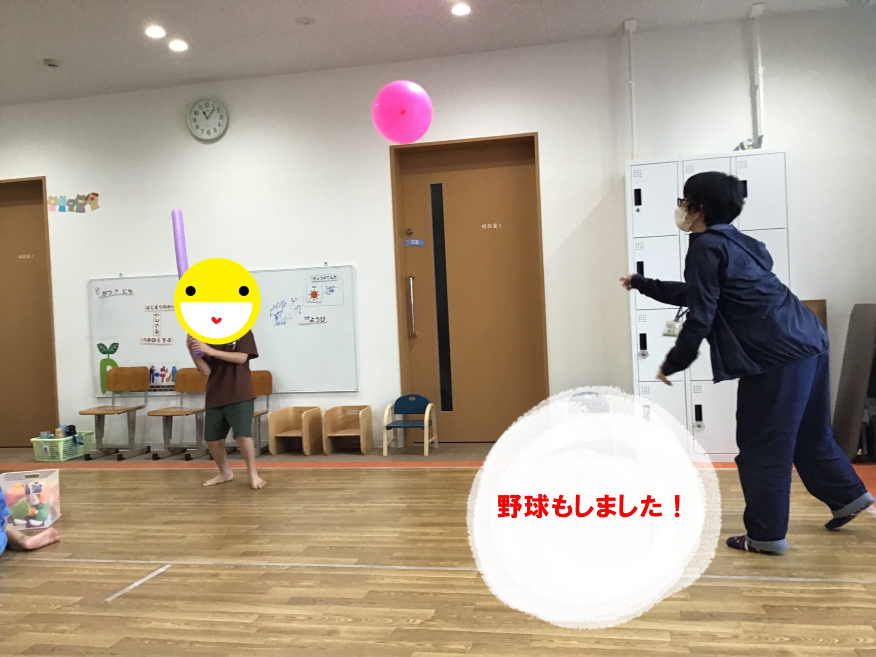Child in a classroom barefoot, holding a purple bat as a pink balloon floats above, with a masked adult nearby and classroom furniture in the background.