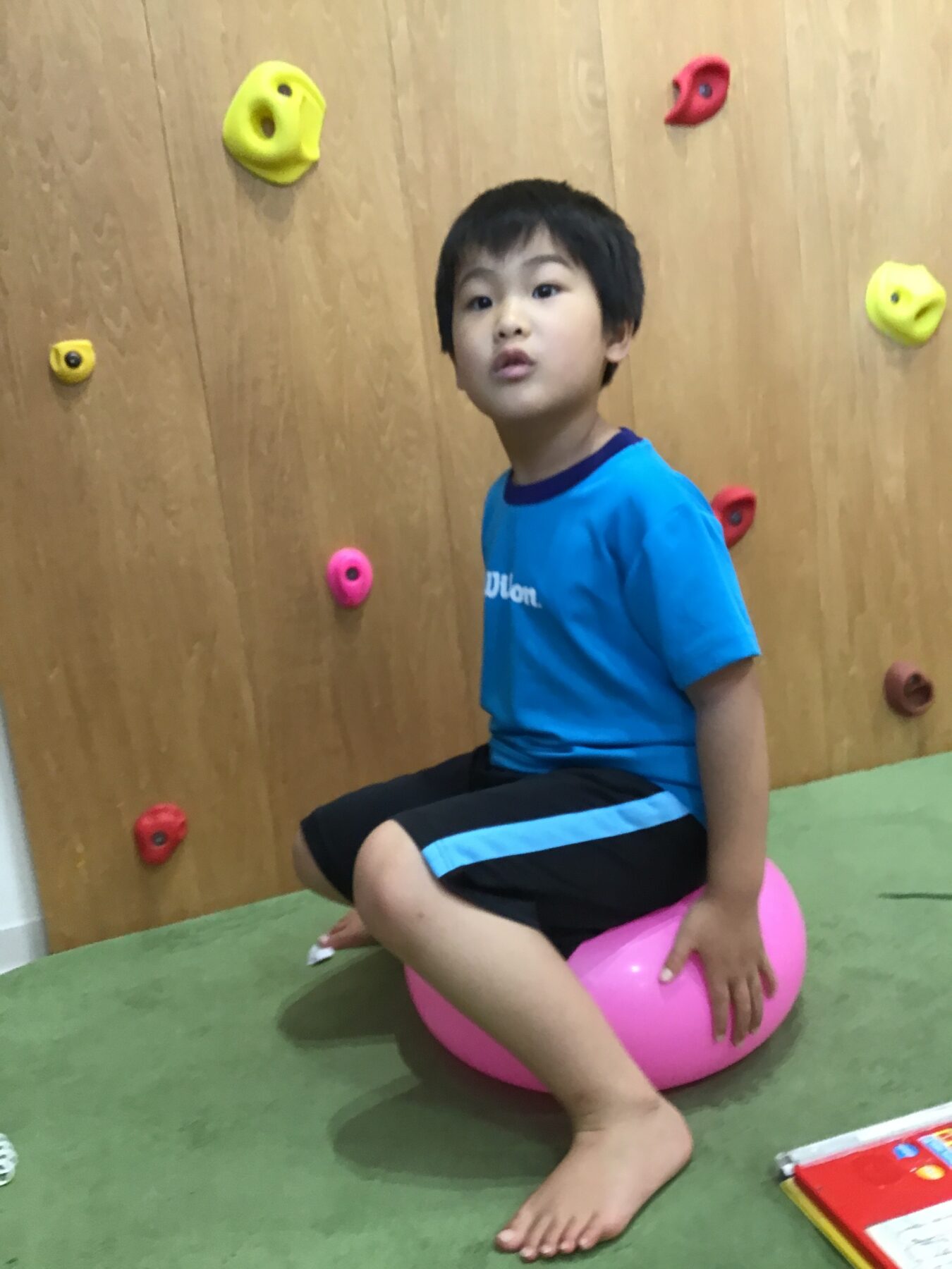 Young boy in a blue shirt sits on a pink exercise ball in a climbing gym with colorful wall holds behind him.