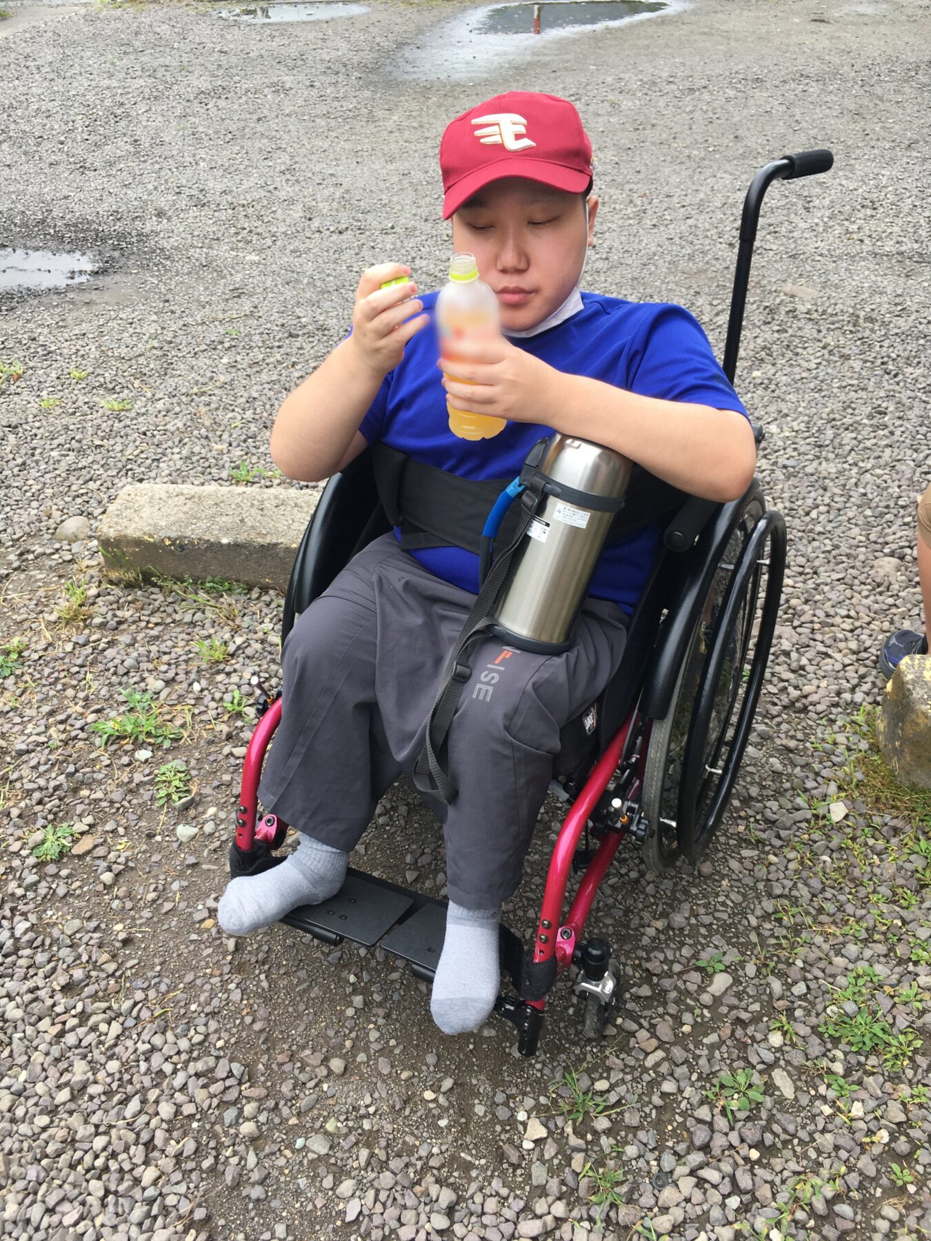 Person in a wheelchair on a gravel surface opens a bottle of orange drink, with a metal bottle holder attached to the chair.