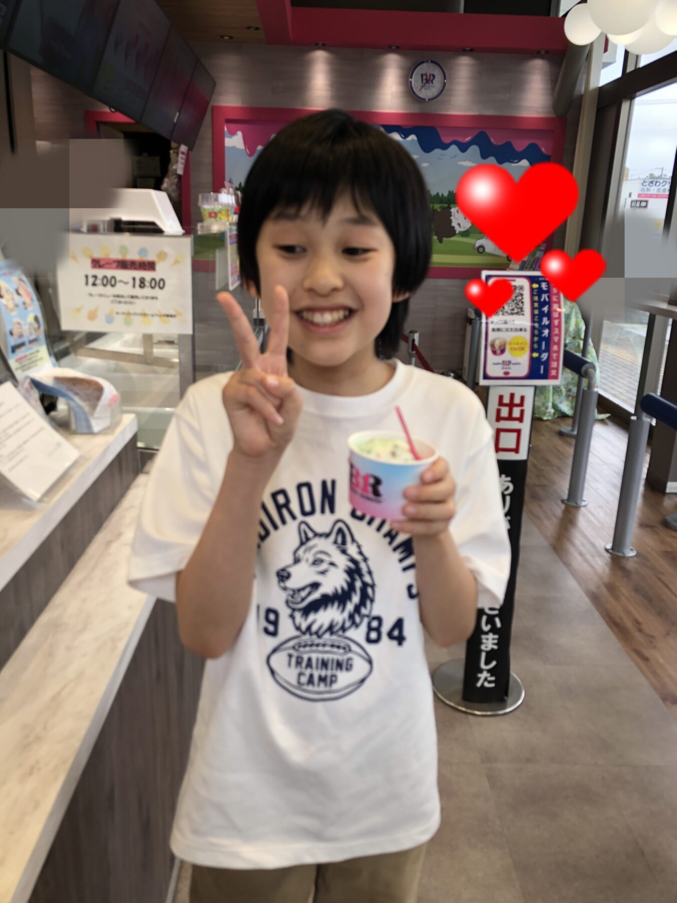 Smiling child in a white T‑shirt makes a peace sign while holding a cup of ice cream in a colorful shop interior.