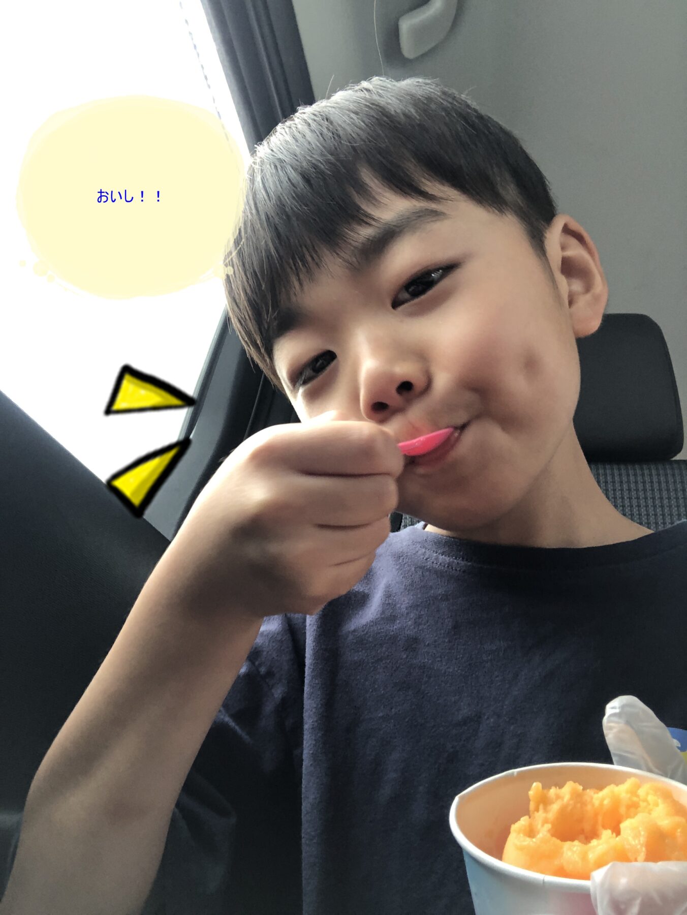 Boy in a car smiling while licking orange ice cream from a cup with a pink spoon; comic-style yellow shapes in the background.