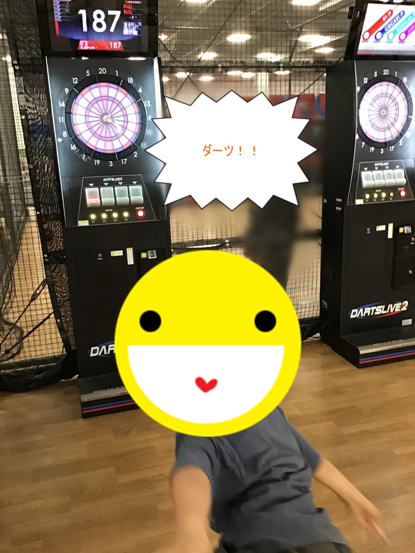 Child seated on the floor in an arcade with two electronic dart machines behind; a large white starburst and big yellow emoji cover the center.