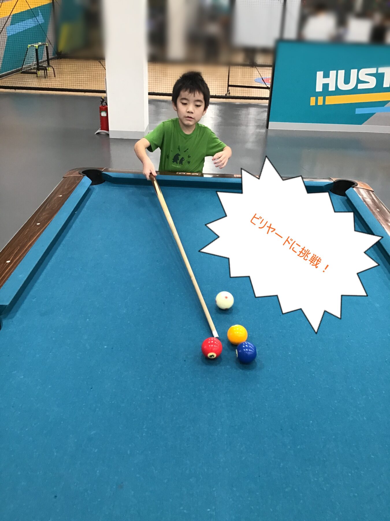 Young boy in a green shirt lines up a pool shot at a blue billiards table, cue poised over red ball and other colored balls nearby.