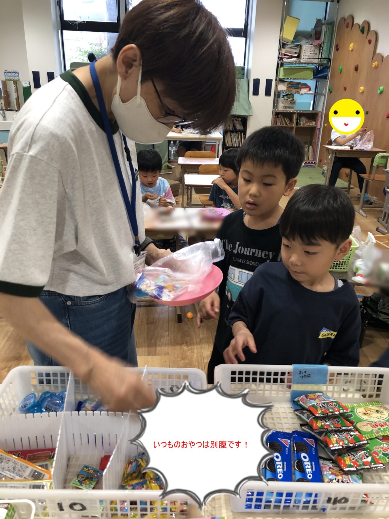 Teacher wearing a mask and lanyard hands out snacks to young students in a classroom shop, with baskets of candy and Oreo packages visible in the foreground.