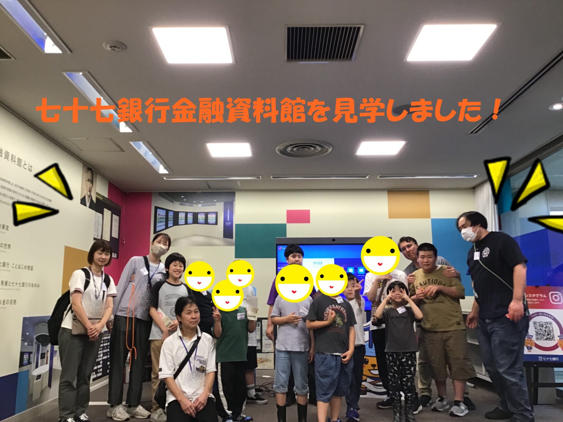 Group of adults and children posing for a photo in a colorful museum-like space, with large orange Japanese text across the top announcing a visit to the 77 Bank Financial Museum.