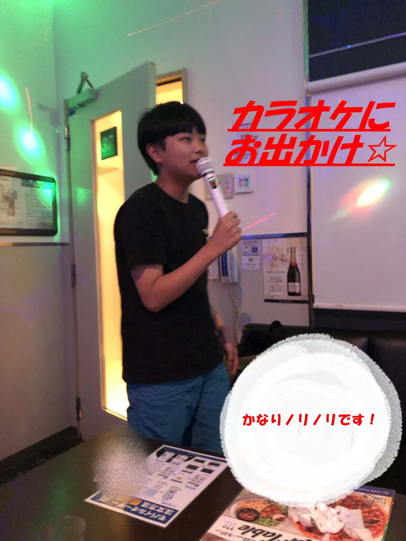 Young man in a black shirt and blue shorts sings into a microphone in a karaoke room with green and pink stage lights.