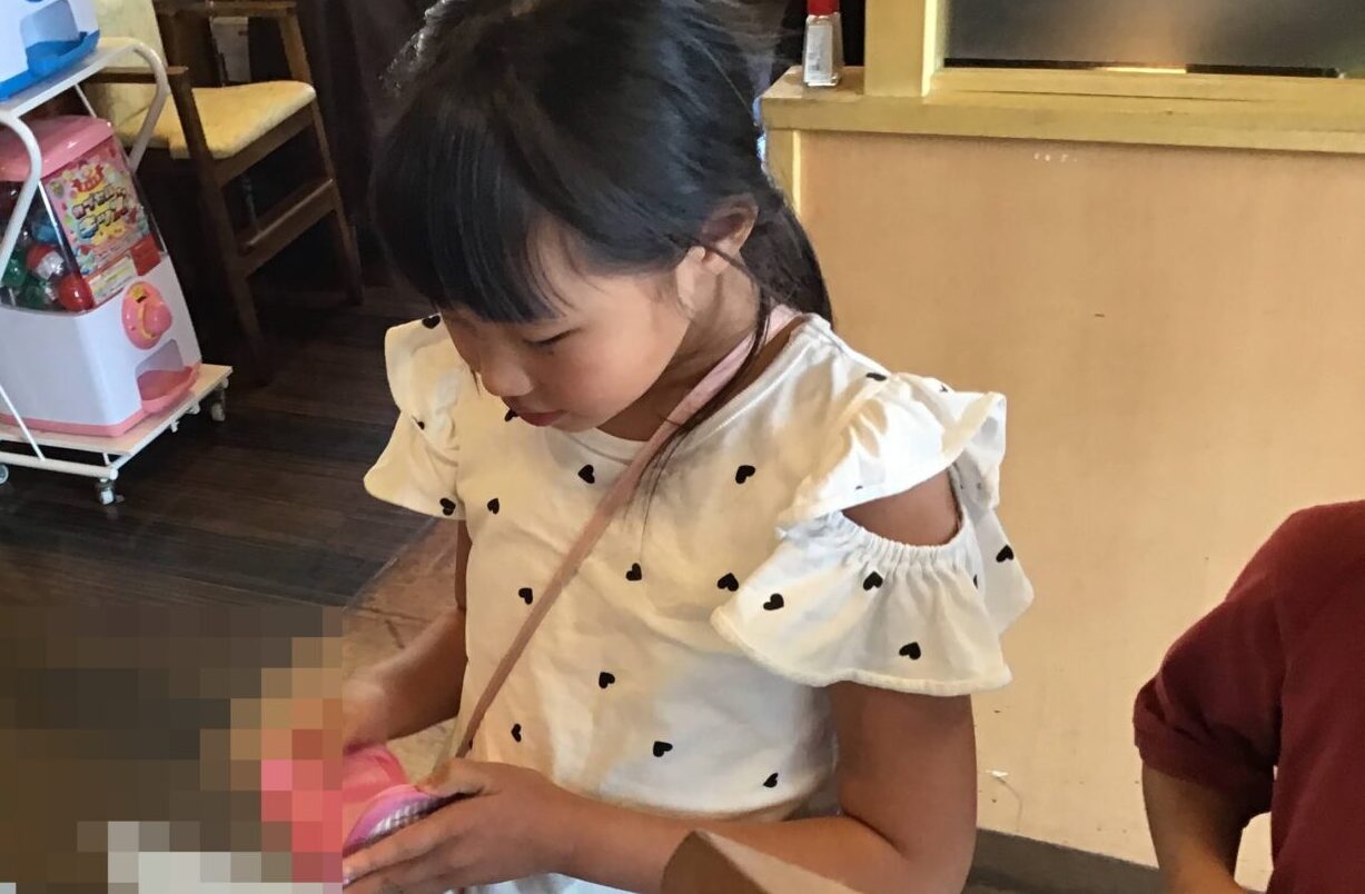Young girl in a white heart-print top stands at a cafe counter, looking down at something in her hands.