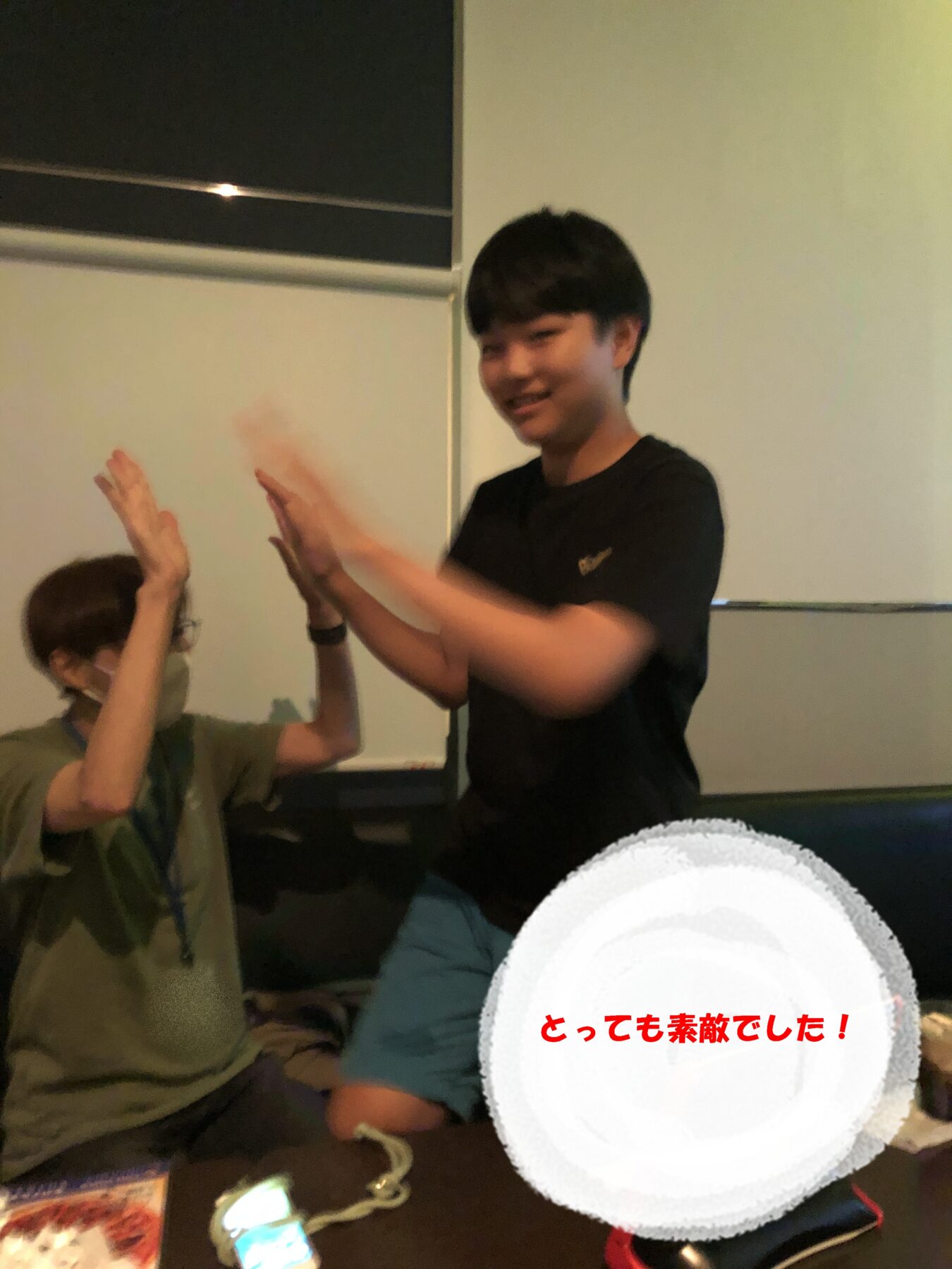 Two people share a high-five indoors: a smiling young man in a black shirt approaches from the right while another person wearing a mask sits nearby on the left.