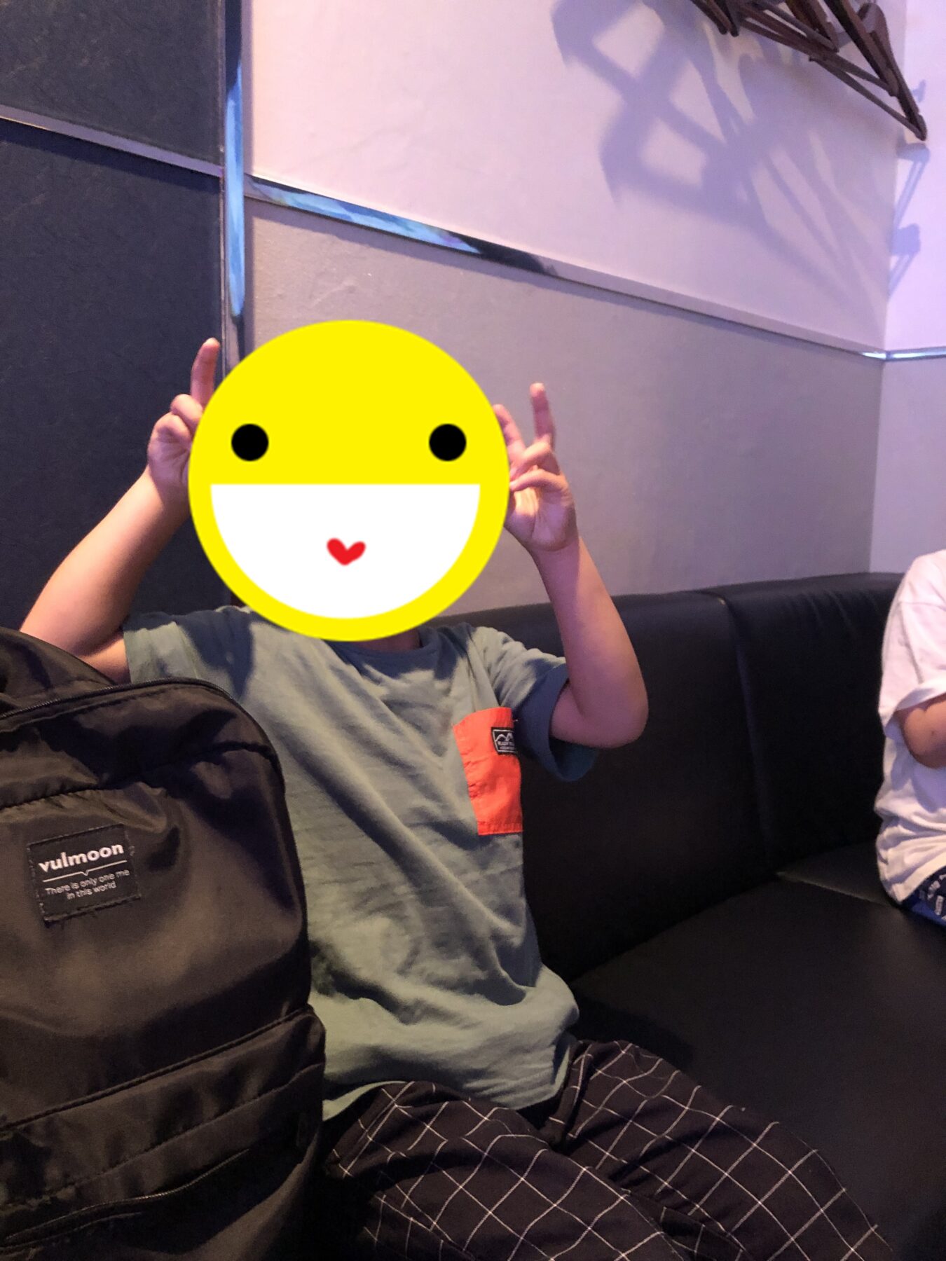 Child sitting on a couch, making peace signs with both hands; a large yellow smiley sticker covers their face, with a black backpack in the foreground