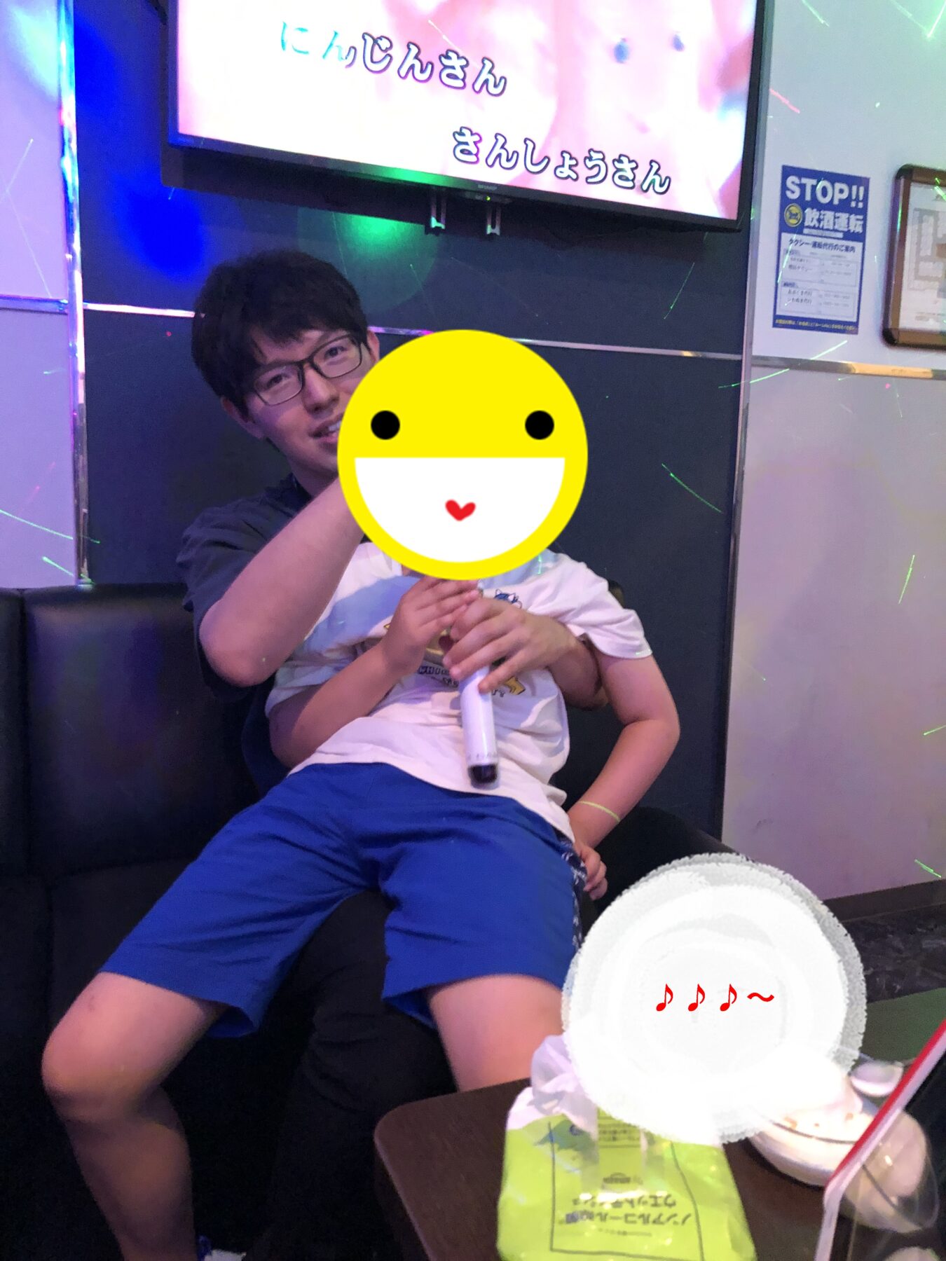 Man with glasses sits on a black couch hugging a child who is holding a microphone; a large yellow smiley sticker covers the child’s face in a karaoke room. Behind them is a screen with colorful text and neon lighting.