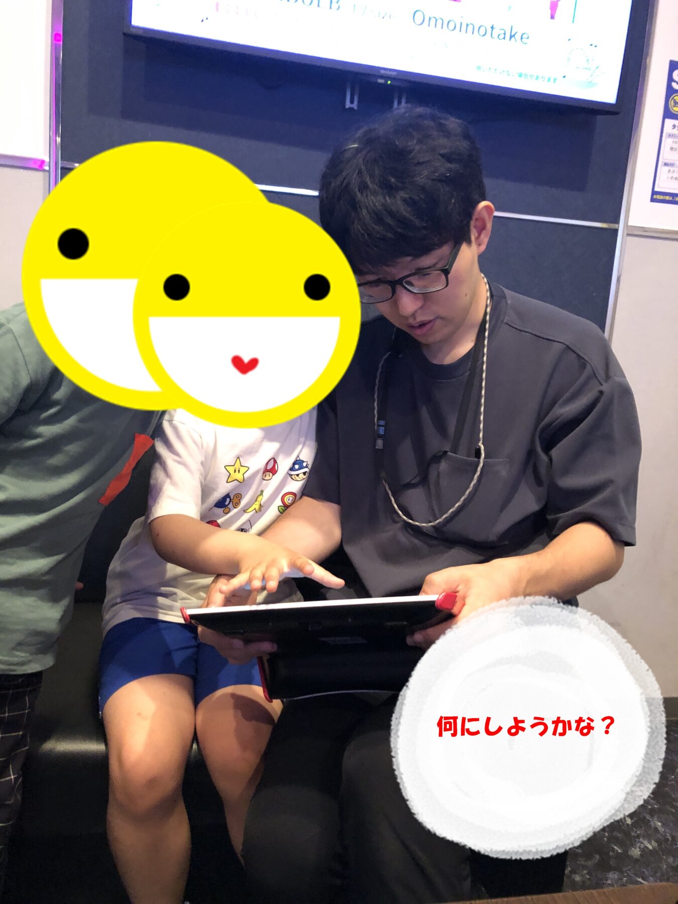 Man with glasses and a child looking at a tablet together; the child's face is covered by a large yellow smiley sticker.