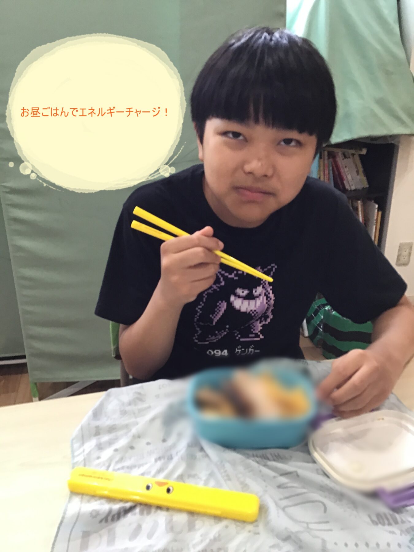 Child sitting at a table holding yellow chopsticks, about to eat from a blue bowl; lunch scene with a speech bubble in Japanese on the left.