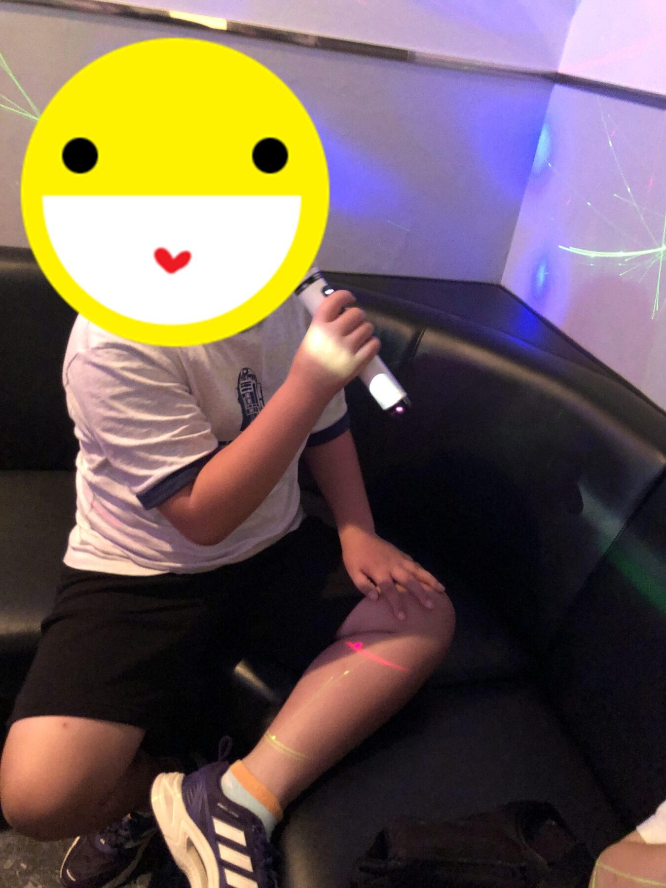 Child sitting on a black couch, holding a white microphone, with a large yellow smiling emoji sticker over their face; colorful laser lights on the wall.