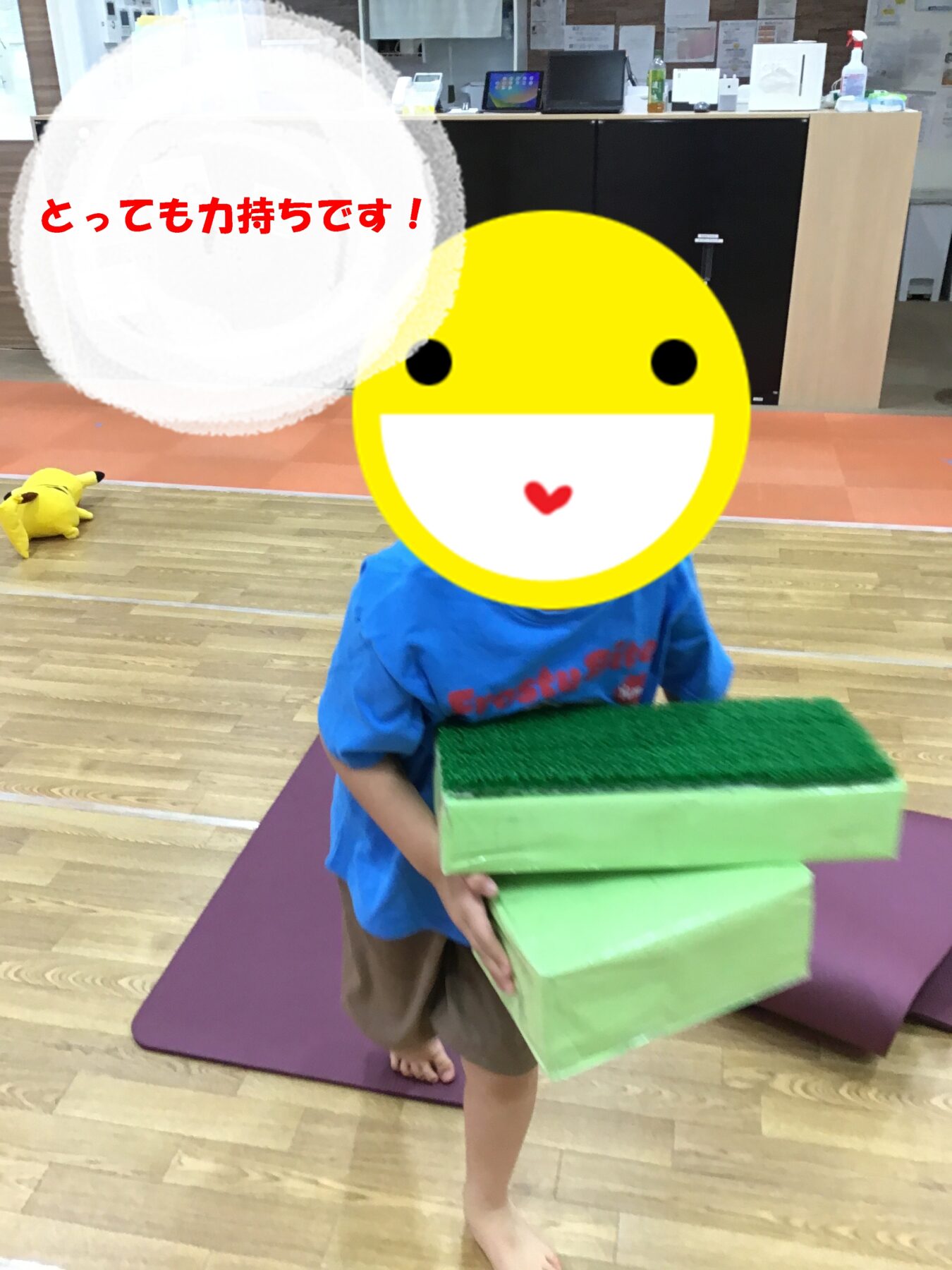 Child wearing a blue shirt carries stacked green foam blocks in a play area; face covered by a large yellow smiley emoji with a speech bubble saying『とっても力持ちです!』(Very strong!).