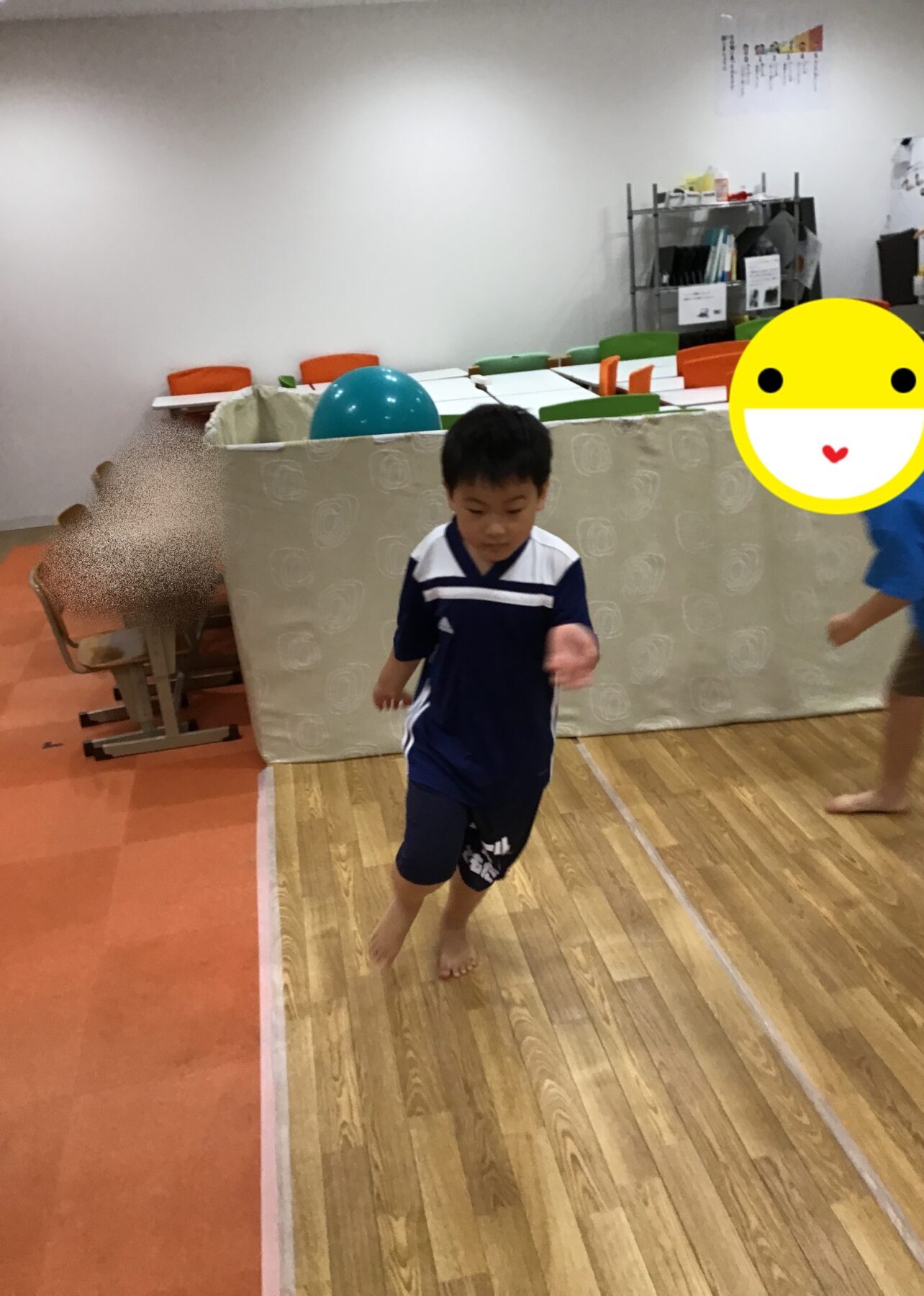 Child in blue sportswear runs barefoot across a wooden gym floor in an indoor play area, with a large yellow emoji covering another person on the right.