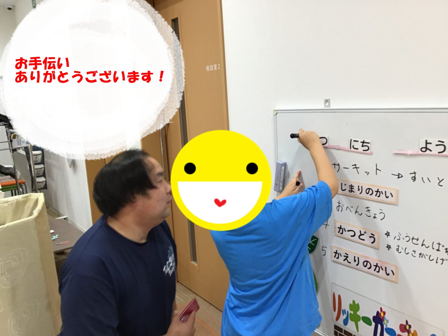 Two people in a hallway work on a whiteboard with Japanese text, one writing as the other watches; warm, collaborative scene.