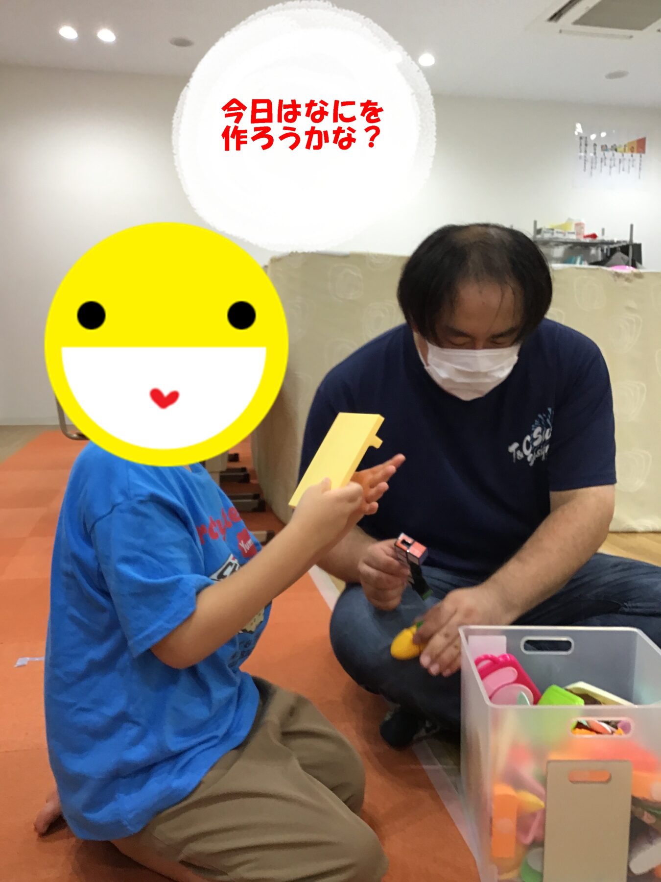 Child wearing a large yellow smiley mask, kneeling on the floor and playing with a toy block beside a masked adult.
