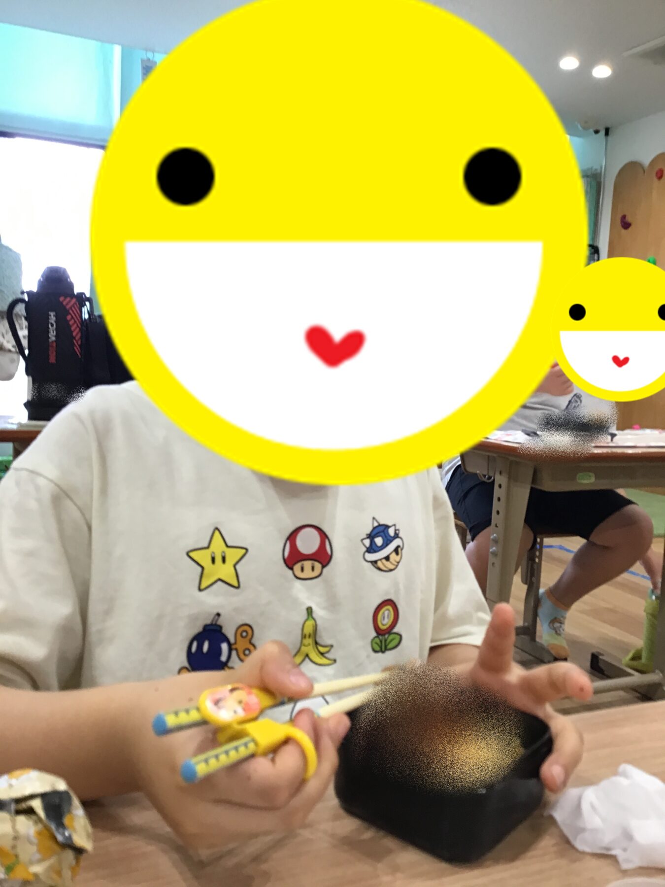 Child sitting at a desk in a classroom, holding colorful pencils; a large yellow emoji covers their face for privacy.