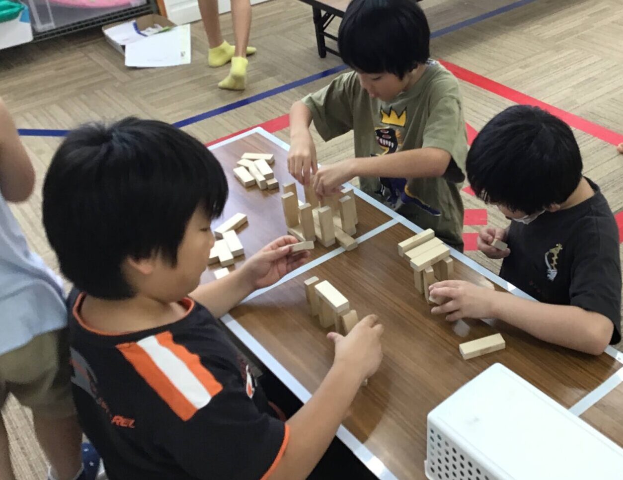 Several children sit around a table building towers with wooden blocks in a classroom, while a girl in a blue dress walks past in the background.