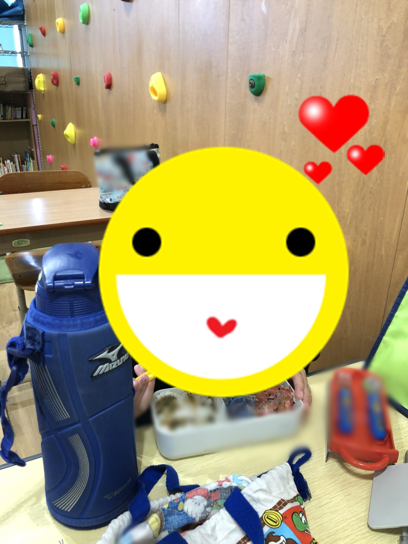 Child with a blue backpack at a table during snack time in a classroom; colorful climbing holds on a wall behind, and a large yellow smiley emoji covers the foreground face.