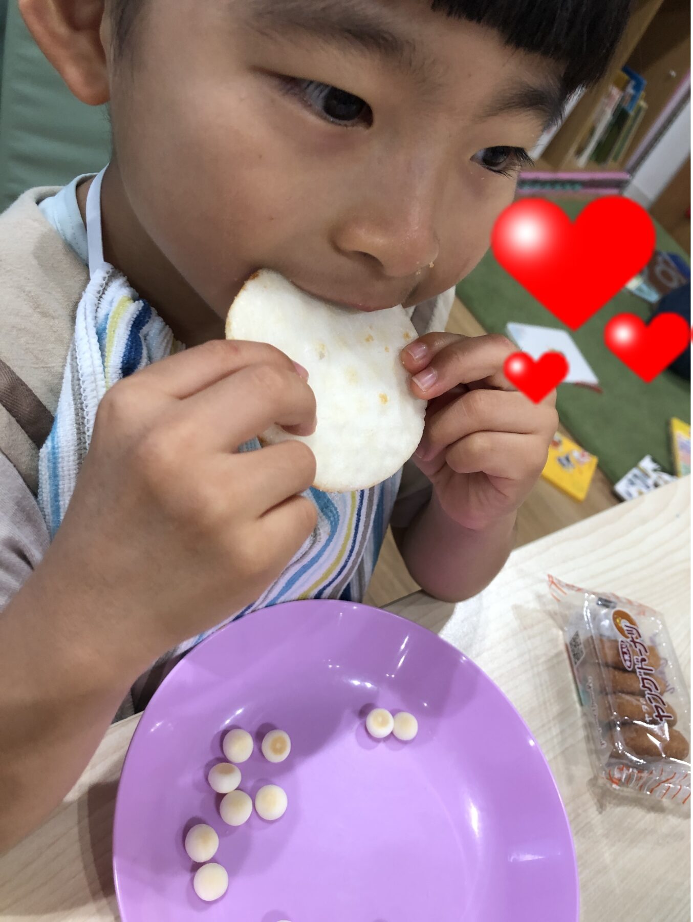 Child biting a white round snack at a table; purple plate with small round crackers nearby, decorative red hearts in the background