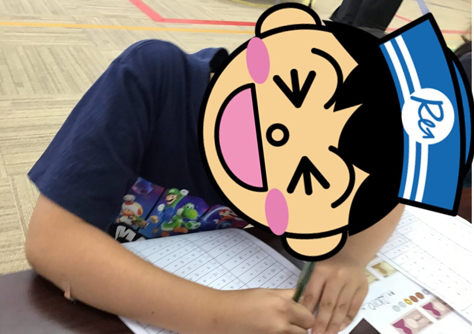 Child in a blue shirt sits at a table, writing on a grid-style worksheet with a pencil; a large cartoon sticker covers their face.