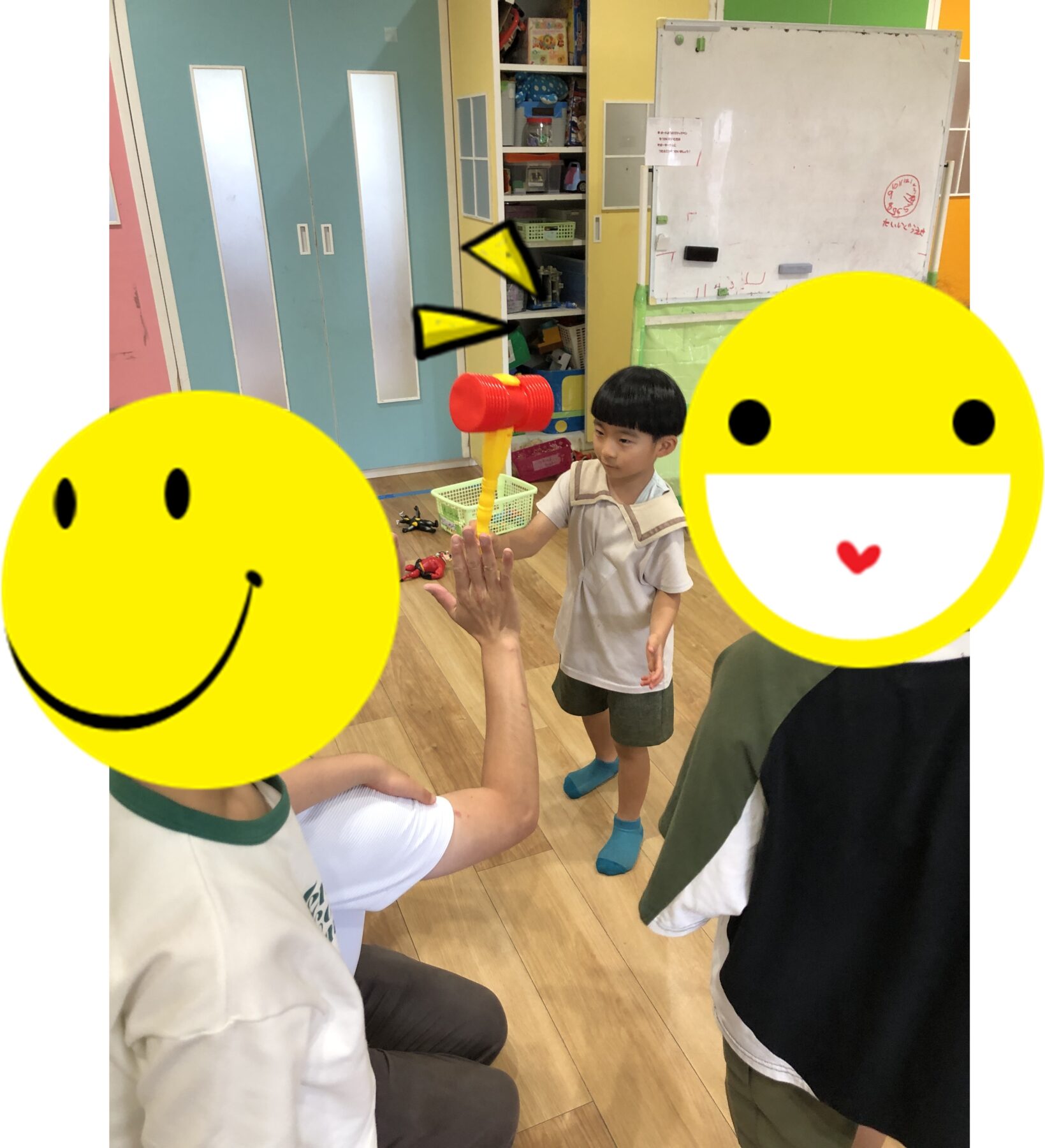 Children in a colorful classroom playfully high-fiving while one holds a red and yellow toy hammer; large smiley-face stickers cover their faces.
