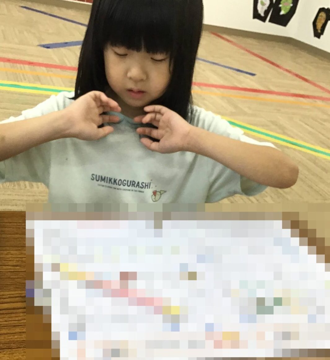 Young child sits at a table with hands raised, focused on activity, papers and a blue pencil case nearby in a classroom setting.