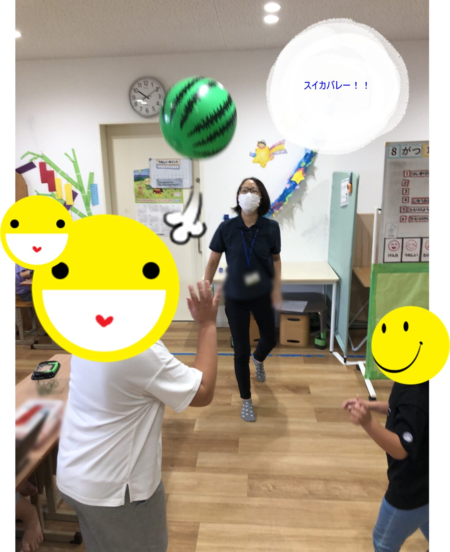 Teacher wearing a mask supervises a watermelon volley game in a classroom; a large green watermelon-ball is midair while kids around are covered with emoji stickers; blue text reads スイカバレー!!