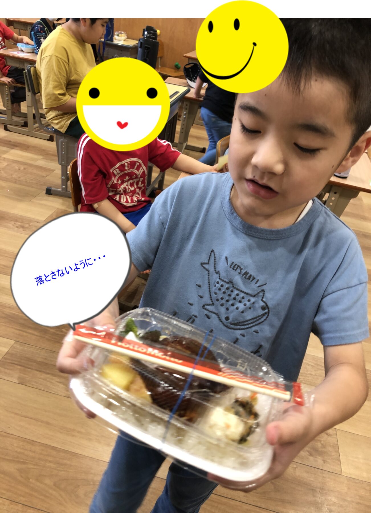 Child in a blue shirt holds a clear plastic bento box with various foods, in a classroom; other children with large yellow smiley overlays in the background; speech bubble shows Japanese text: 落とさないように…